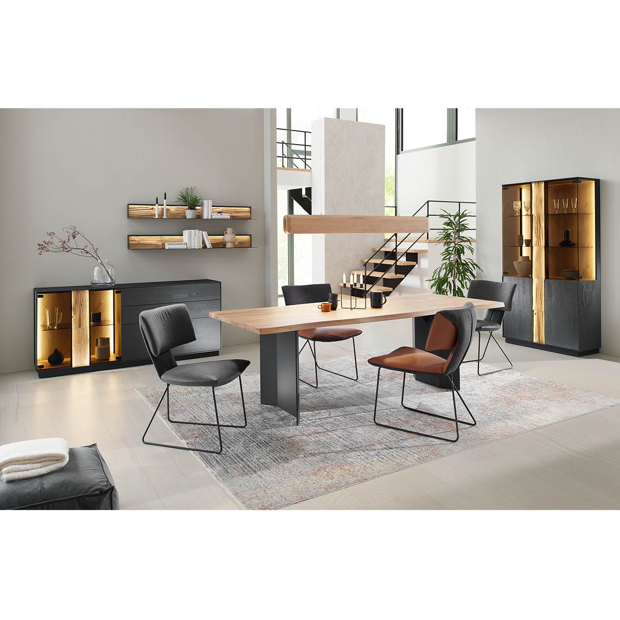 Modernes Esszimmer mit rechteckigem Holztisch umgeben vor drei Stühlen auf filigranem Metallgestell. Darüber hängend eine Hängeleuchte aus massivem Holzbalken an Stahlseil. Im Hintergrund die passenden Esszimmermöbel.