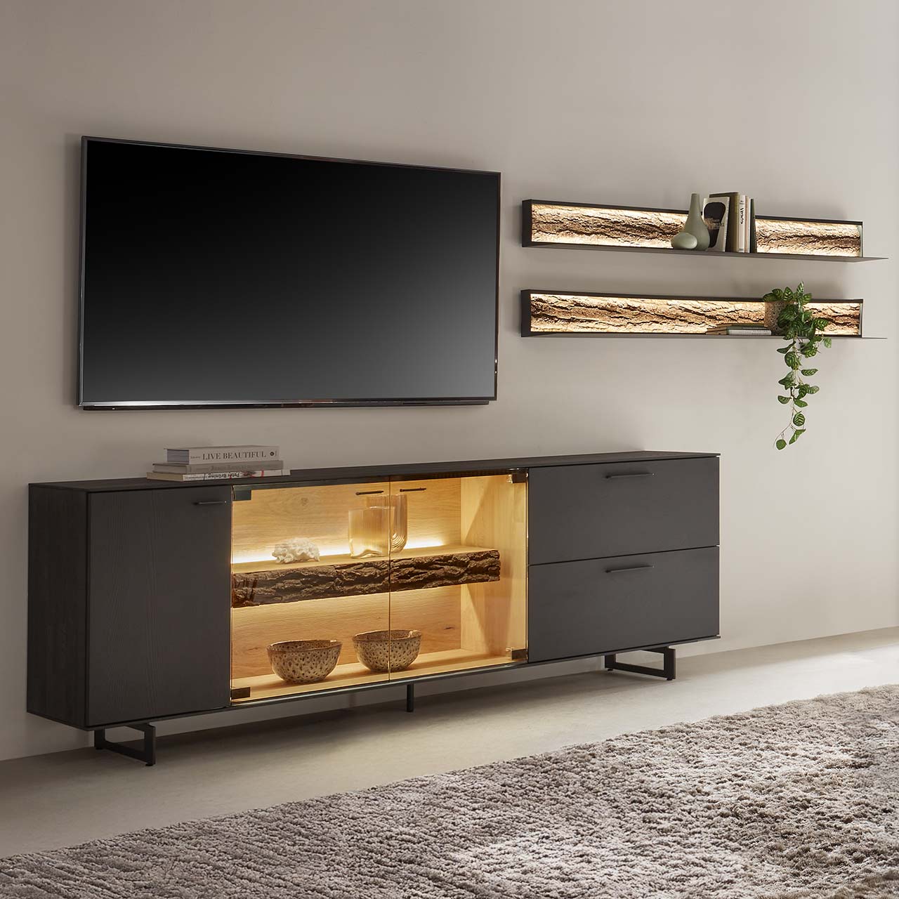 Modernes Wohnzimmer mit vierteiliger Wohnwand bestehend aus einem Lowboard aus massiver Kerneiche Natur, anthrazit gebeizt mit beleuchtetem Glasausschnitt auf Metallkufen und zwei Wandregale aus Metall mit sichtbarer Baumrinde
