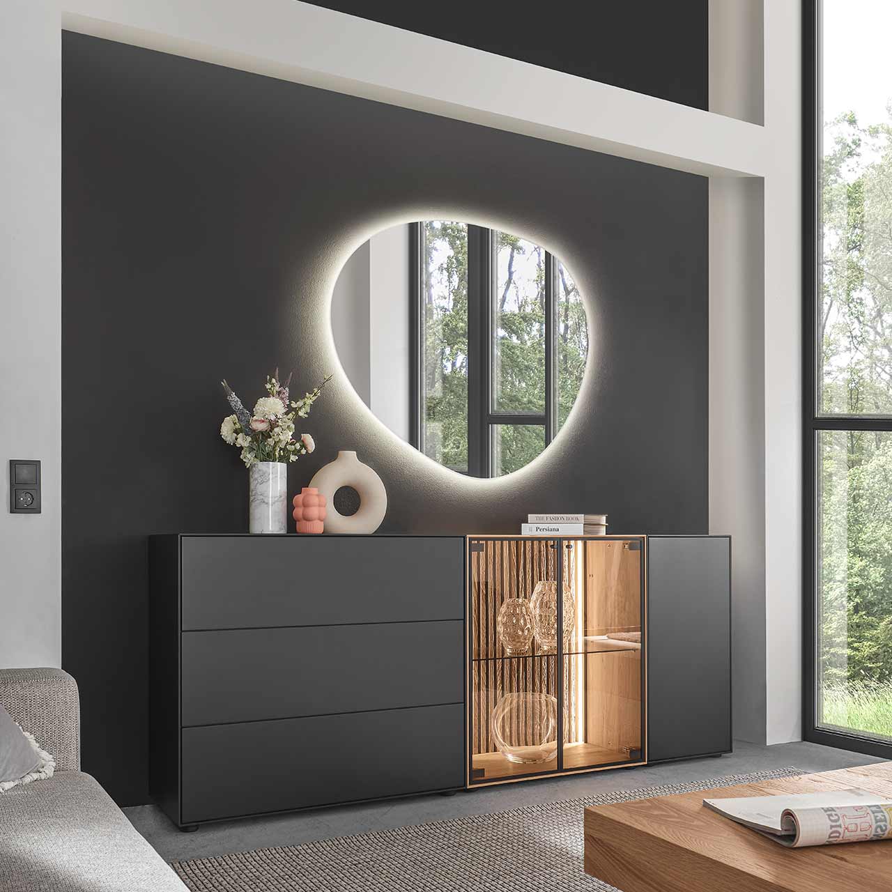 Modernes Sideboard aus massiver Birke anthrazit mit Holztür, zwei Glastüren, beleuchteter heller Holz-Lamellenrückwand und Glaseinlegeboden sowie zwei Schubkästen und Klappe in Anthrazit. Darüber hängt ein asymmetrischer Spiegel - vor dunkelgrauer Wand.