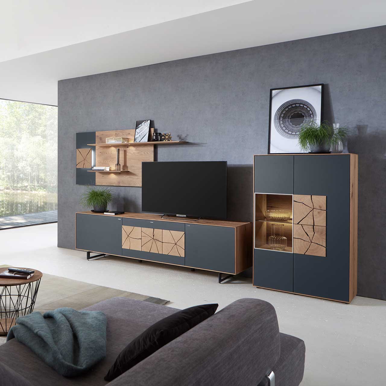 Milieubild Hartmann CAYA - Standelement 7170-7094A re. Wohnzimmer mit moderner dreiteiliger Massivholz Wohnwand mit Mattglas Anthrazit Fronten und dekorativen Hirnholzscheiben: Langes Lowboard mit Hirnholz und Kufen, Wandpaneel mit Regalböden, Highboard mit beleuchtetem Glasausschnitt und Hirnholz.