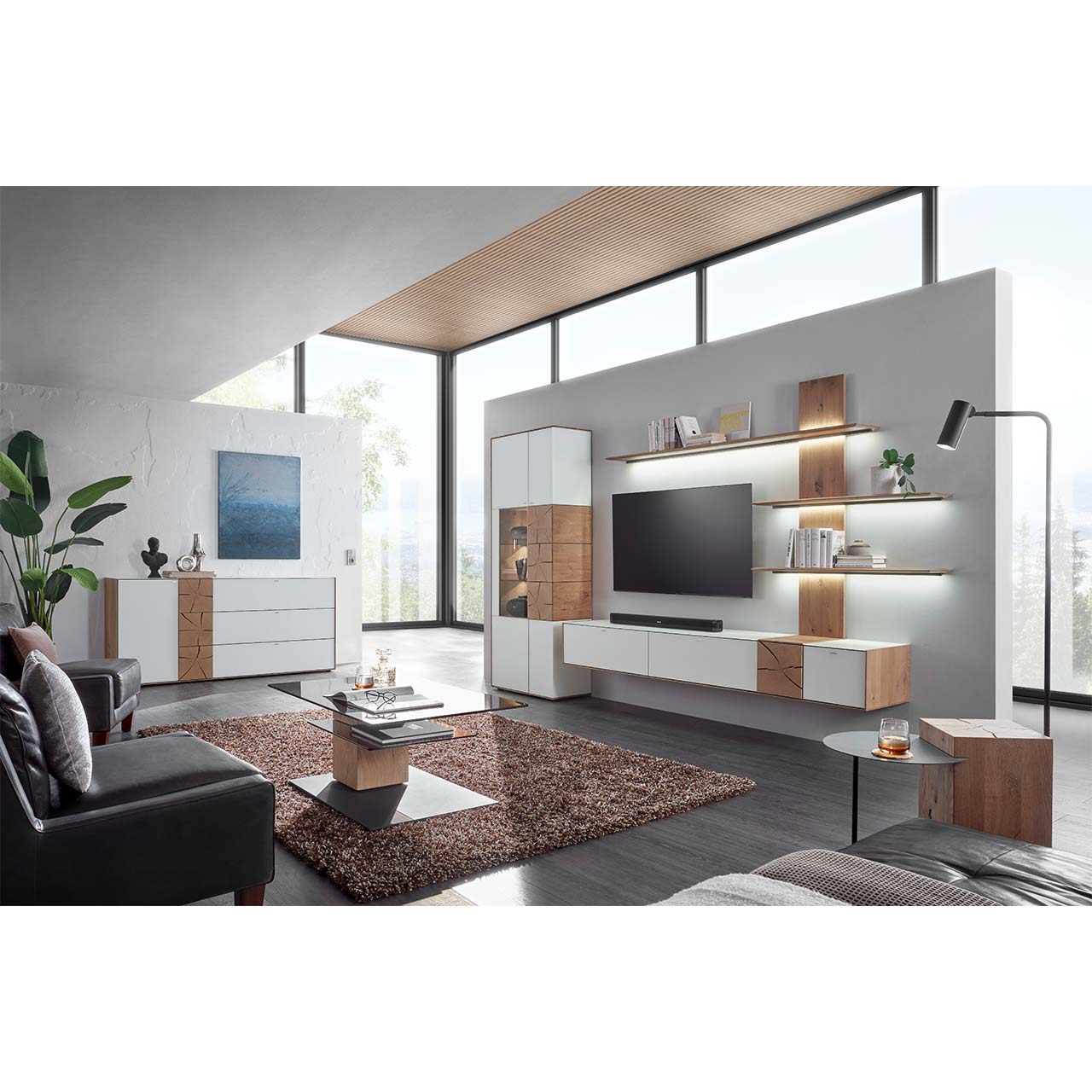 Wohnzimmer mit TV und moderner Wohnwand aus massiver Kerneiche mit Mattglas weißen Fronten, mit Hängelowboard, Wandpaneel mit Regalböden und Standvitrine. Links das passende Sideboard. Im Vordergrund ein Couchtisch mit rechteckiger Glasplatte.