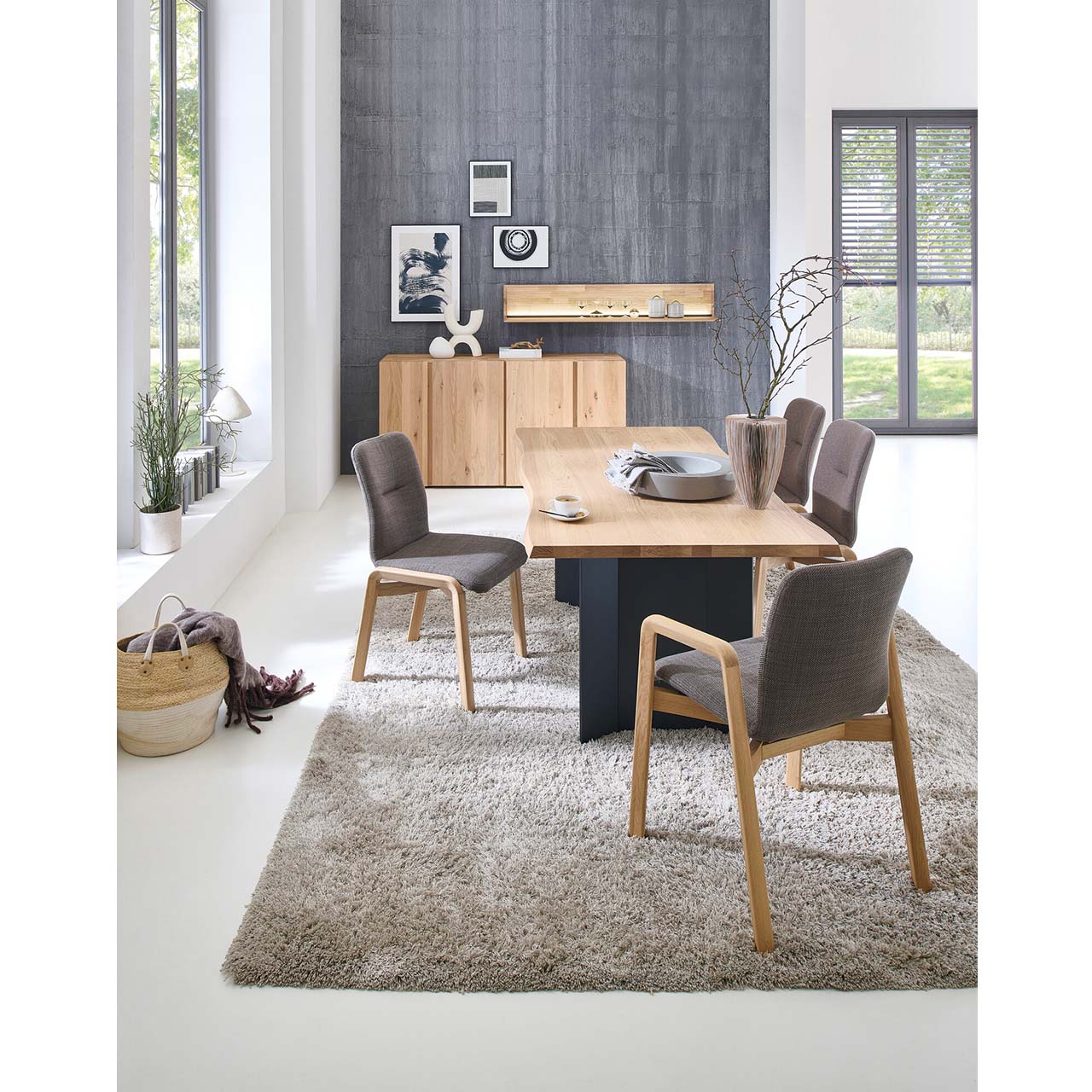 Modernes Esszimmer mit massivem Eichenholztisch mit rechteckiger Tischplatte und Untergestell in Anthrazit, umgeben von vier klassischen Stühlen mit Holzgestell, Lehnen und hellem Stoffbezug. Hinten an der Wand passendes Sideboard mit Wandpaneel. 
