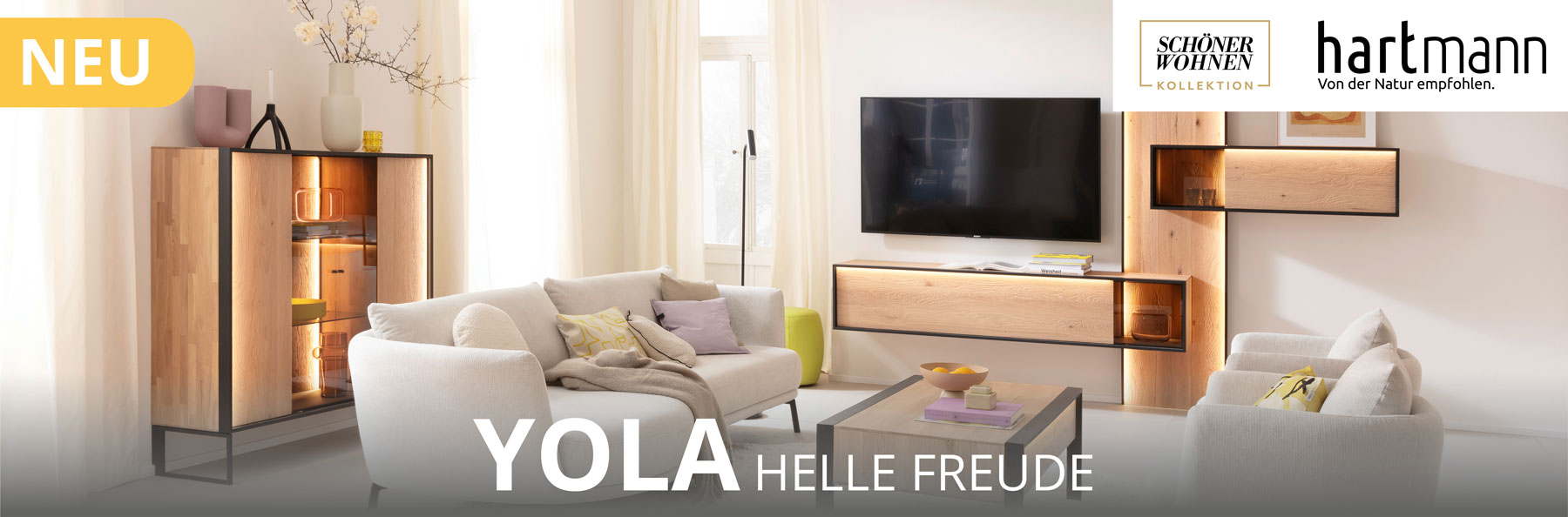 Möbelserie YOLA hell und freundlich: Wohnzimmermöbel aus massiver Kerneiche Natur sandgestrahlt kombiniert mit Metall anthrazit. Ambientebild zeigt beleuchtetes Wandpaneel mit Hängevitrine und Lowboard und Highboard mit Glasfront auf Metallkufen.