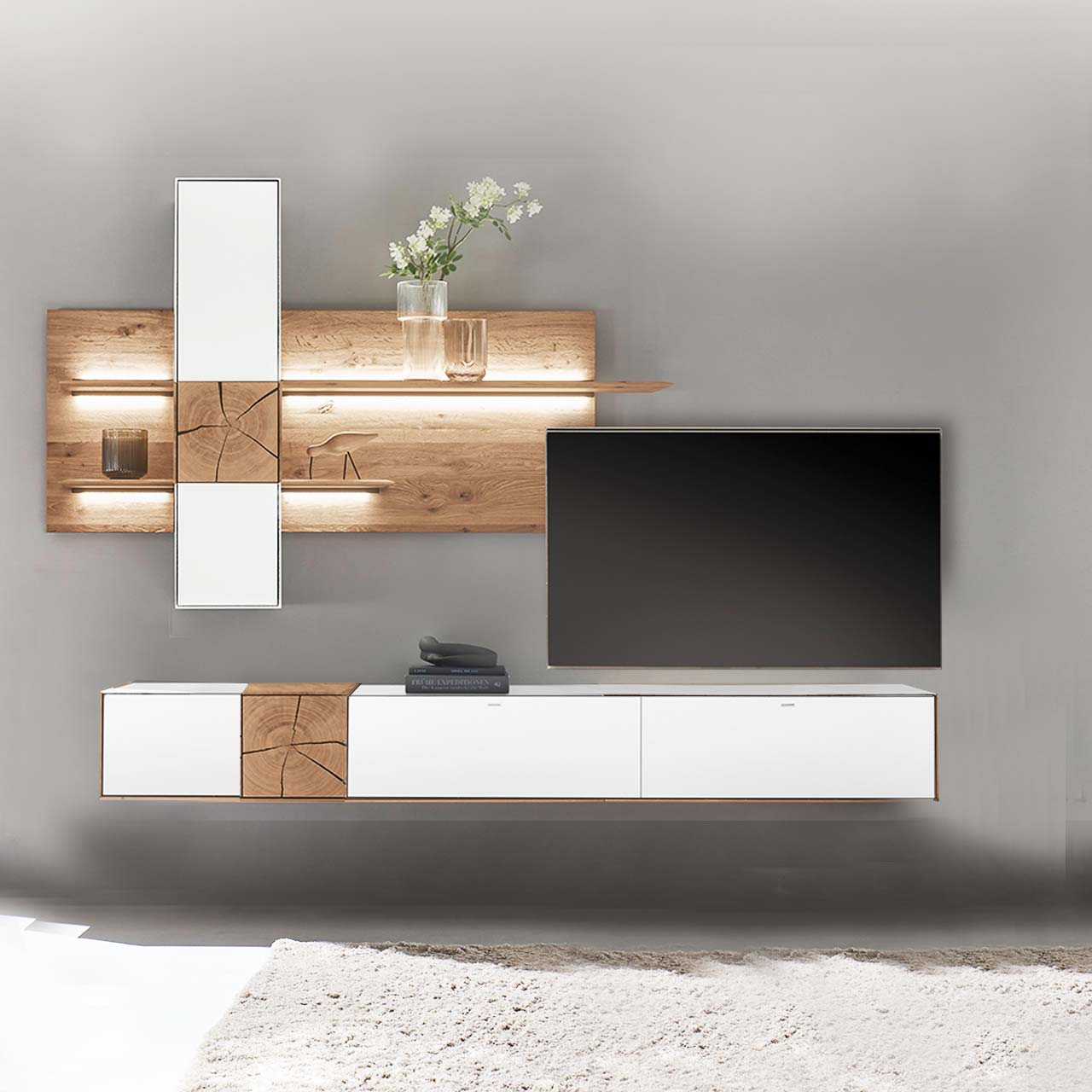 Moderne Wohnwand aus massiver Kerneiche mit Mattglas weiß Fronten. Unten ein langes Hängelowboard mit dekorativen Hirnholzscheiben, oben eine Wandpaneel-Kombination mit Regalböden, Schrank und indirekter Beleuchtung, dazwischen hängender Fernseher.