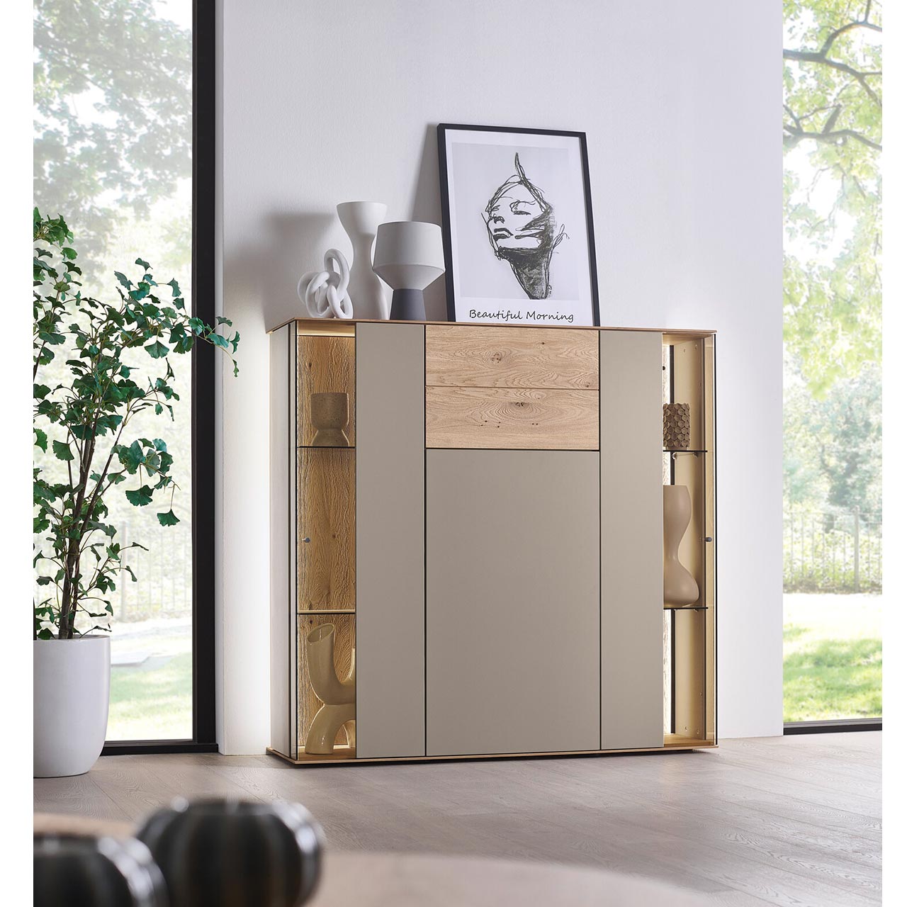 Hartmann SVEA - Highboard 8660-7141 - inkl. Beleuchtung Modernes Highboard aus massiver Kerneiche Natur mit Lackfronten in Sand und Riffholz. Mit einer Lacktür unten, einem Riffholz-Schubkasten oben und jeweils rechts und links einer Glas-Lacktür mit beleuchteter Riffholzrückwand - in Frontansicht.