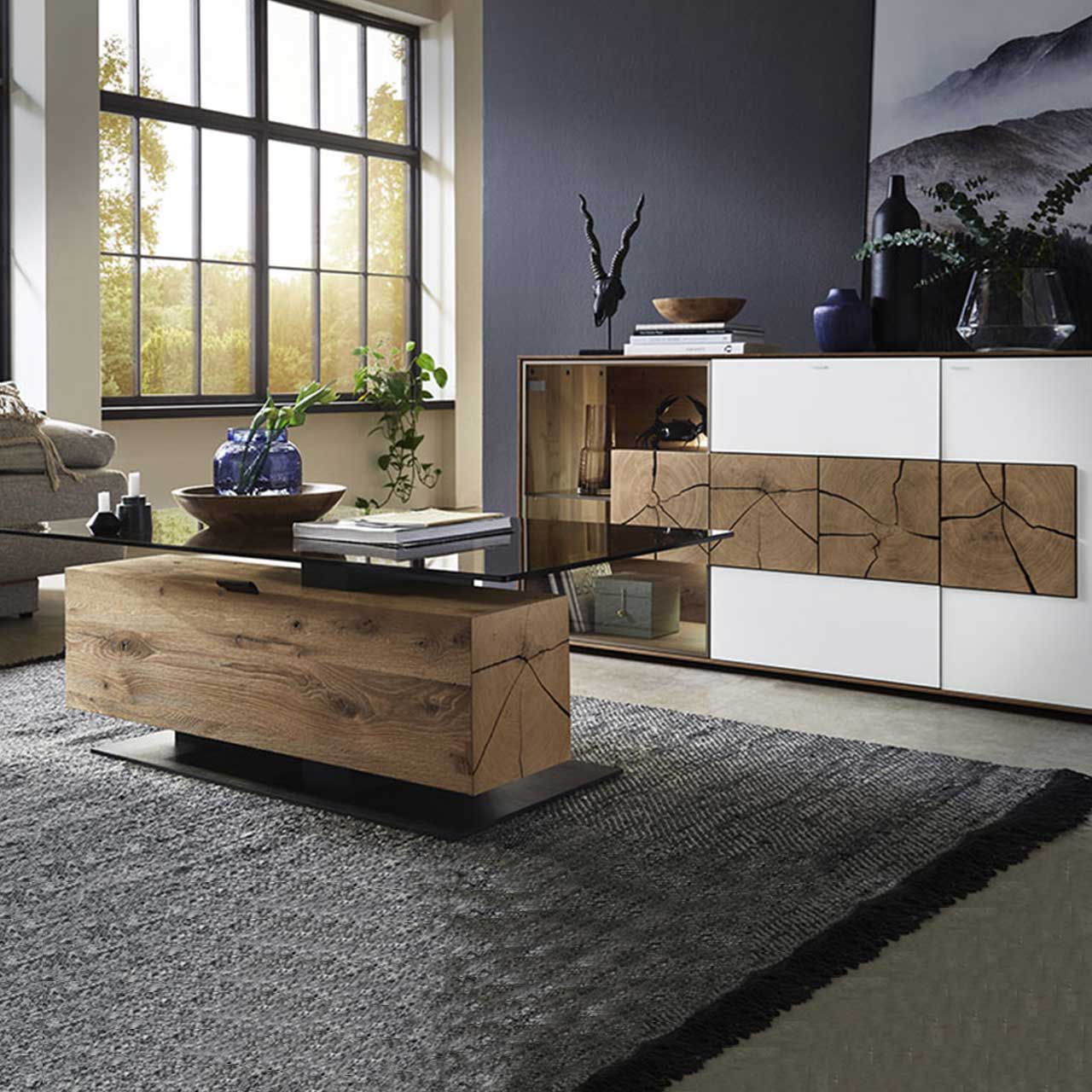 Bildausschnitt eines Wohnzimmers mit modernem Lowboard aus massiver Kerneiche Umato mit weißfarbenen Mattglasfronten, drei rechteckigen Hirnholzscheiben und Glastür mit beleuchtetem Glasboden und Hirnholzblock, davor passender Couchtisch.