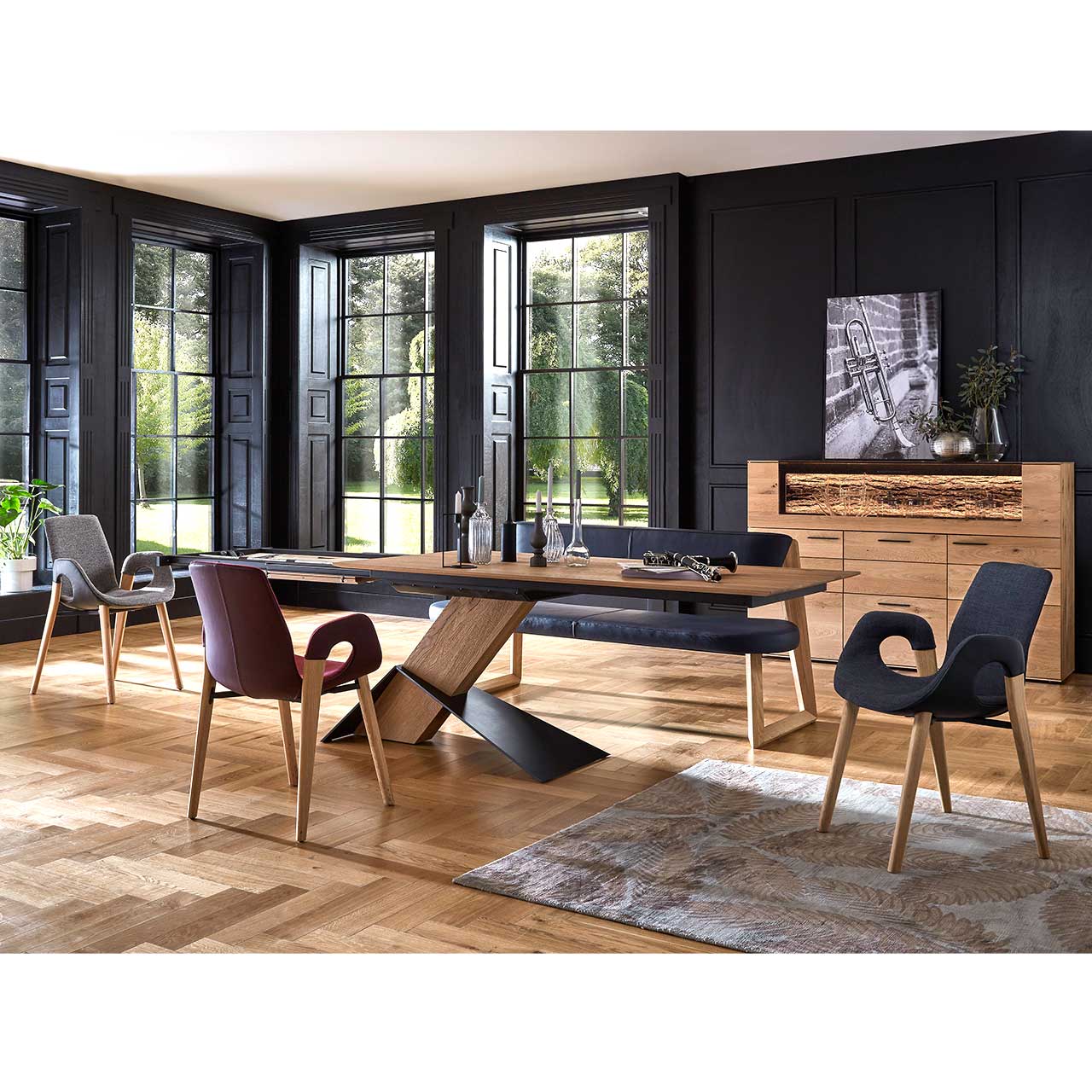 Modernes Esszimmer mit massivem, rechteckigem Holztisch, drei Stühlen auf Holzgestell und einer Sitzbank in schwarzem Leder. Dahinter vor dunkler Wand das passende Highboard mit Glasausschnitt.