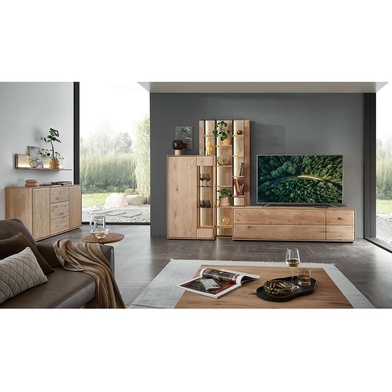 Modernes Wohnzimmer mit dreiteiliger Massivholzwohnwand bestehend aus einem Highboard mit Glastür, einem Hochregal mit Holzböden, beleuchteten Glasseiten und geschroppter Rückwand und einem Lowboard. Davor eckiger Couchtisch, links passendes Sideboard.