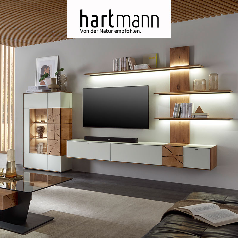 Hartmann CAYA - Massivholzmöbel aus Kerneiche Umato mit Mattglas weiß oder anthrazit Fronten und dekorativen Hirnholzscheiben. Ambientebild zeigt Hängelowboard weiß, beleuchtetes Wandpaneel aus Holz und Highboard mit Glastüren und Hirnholzscheiben.