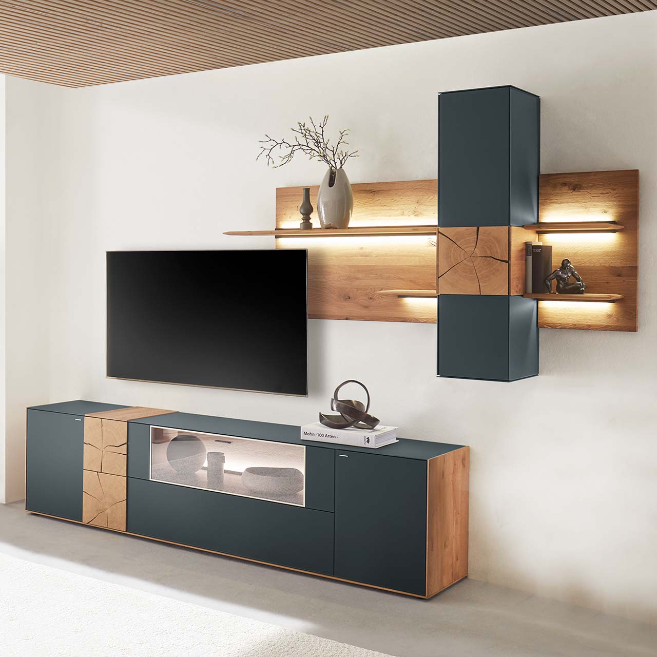 Moderne Wohnwand aus massiver Kerneiche mit anthrazitfarbenen Fronten. Unten ein Lowboard mit Vitrine und dekorativer Holzapplikation, oben eine Wandpaneel-Kombination mit Regalböden, Schrank und indirekter Beleuchtung, dazwischen hängender Fernseher.
