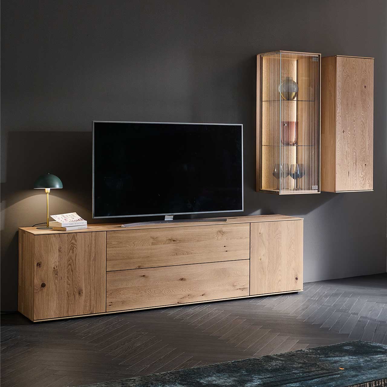 LINEA Leonardo Living - Wohnwand 8540 Nr. V22 Wohnzimmer mit Fernseher und moderner dreiteiliger Wohnwand aus Kerneiche Natur: Ein schlichtes Lowboard, eine Hängevitrine mit satiniertem Glas und ein Hängeschrank – platziert vor einer dunkelgrauen Wand.
