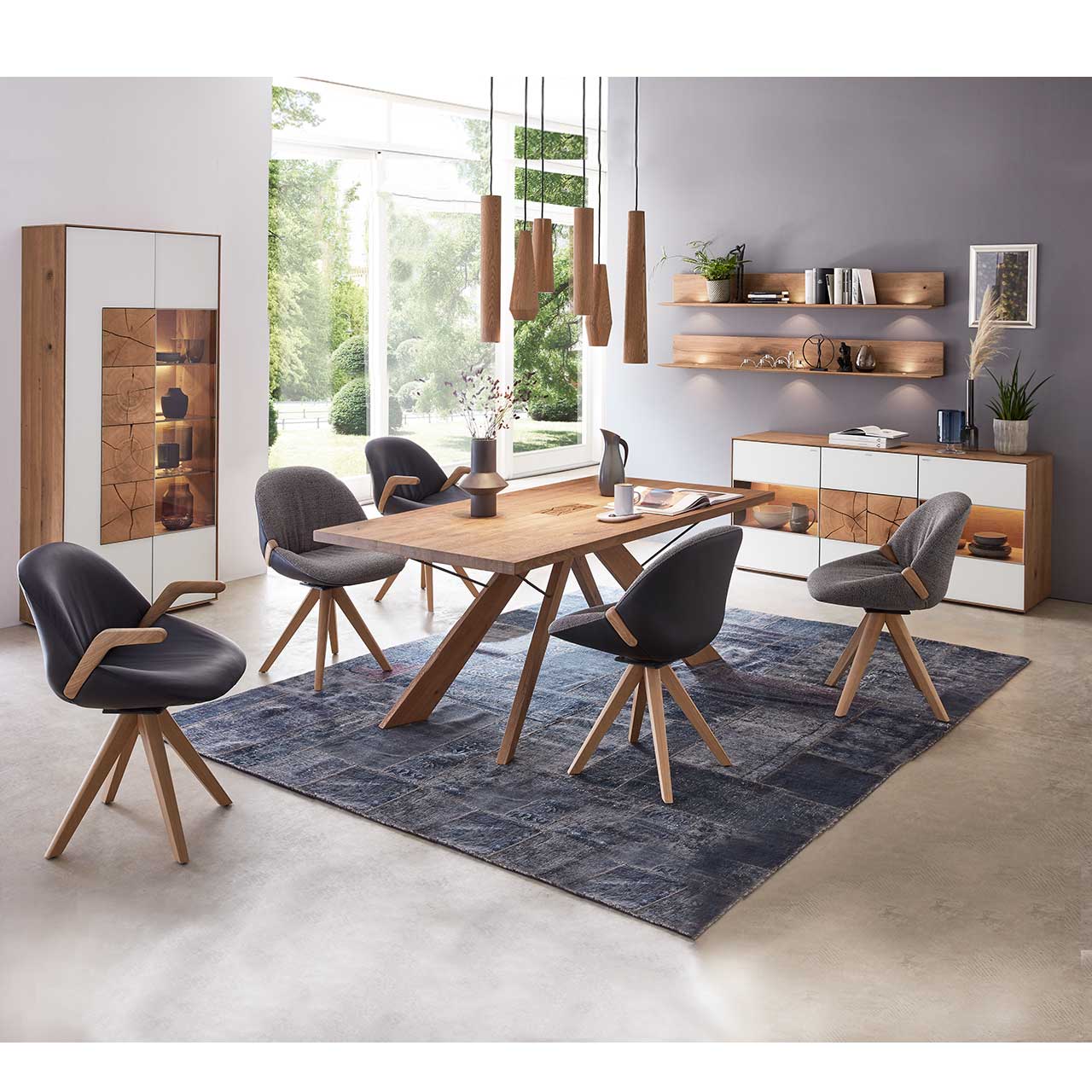 Milieubild Esszimmer - Hartmann CAYA - Standvitrine 7170-0096W li. Esszimmer mit massivem Holztisch und fünf Stühlen mit Holzgestell, links an der Wand stehend eine Strandvitrine in Mattglas weiß und beleuchtetem Glasausschnitt mit drei Glasböden. Rechts das passende Sideboard und zwei Wandregale.