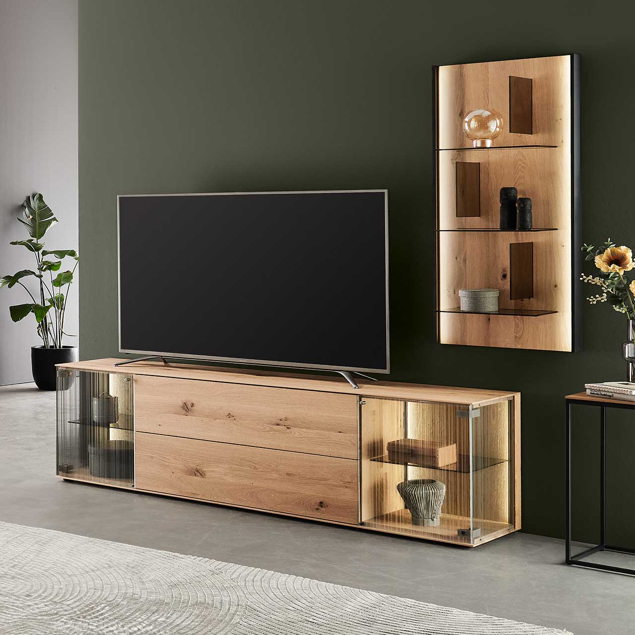 LINEA Leonardo Living - Wohnwand 8540 Nr. V26 Modernes Wohnzimmer mit Fernseher und zweiteiliger Wohnwand aus Kerneiche Natur: Lowboard mit satinierten Glastüren und Holzschubkasten und Wandpaneel mit drei Glasböden – platziert vor einer dunkelgrünen Wand.