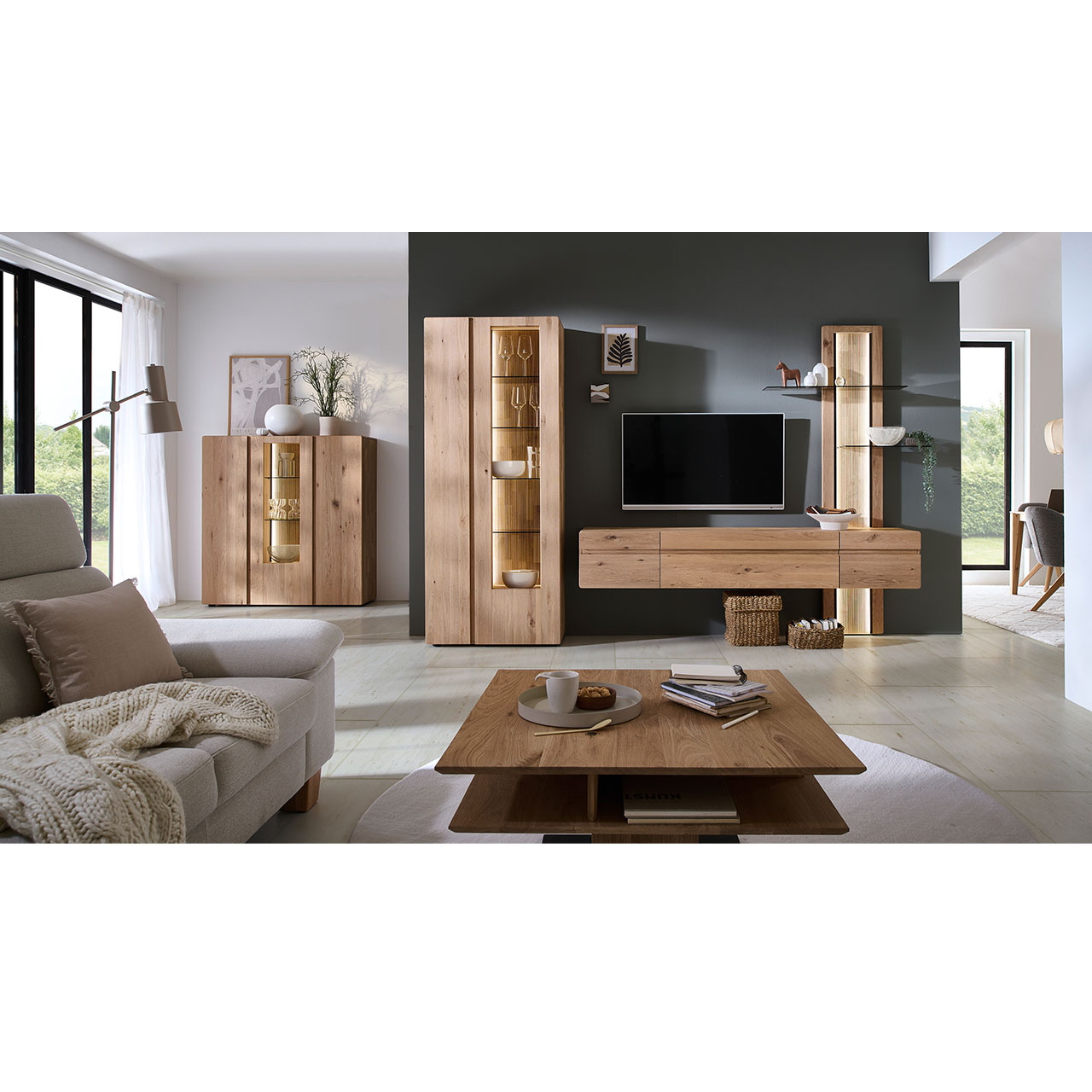 Milieubild - Hartmann VIDA - Wohnkombination 8630 Nr. V26 - zweiteilig - Kerneiche Natur Modernes Wohnzimmer mit dreiteiliger Wohnwand aus massiver Kerneiche Natur: Standvitrine mit zwei Türen, Hängelowboard und Aufsatzpaneel mit beleuchteten Glasböden sowie geschroppter Rückwand. Links hinten an der Wand passendes Highboard.