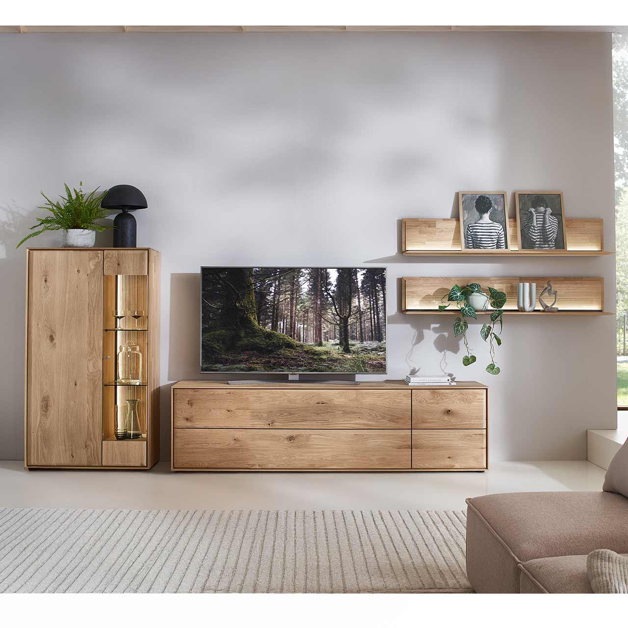 Moderne vierteilige Wohnwand aus massiver Kerneiche Natur bestehend aus einem Lowboard mit Schubkästen, einem Highboard mit Glastür sowie zwei beleuchteten Wandpaneelen mit Holzboden und geschroppter Rückwand.