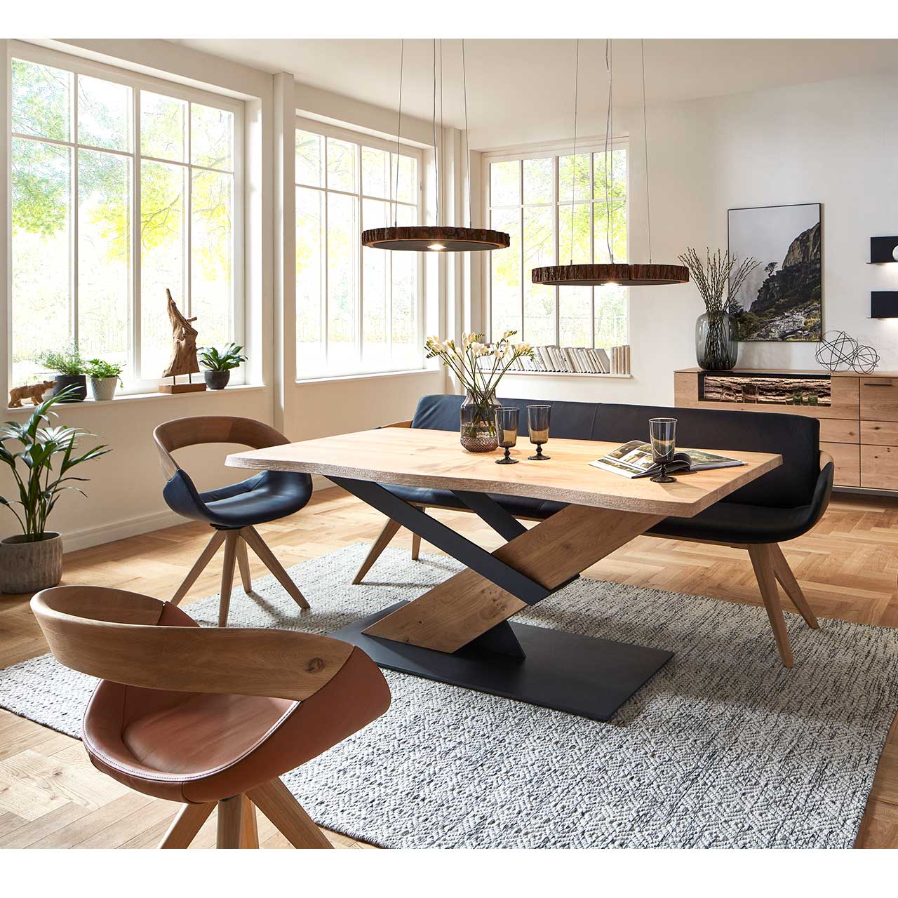 Modernes Esszimmer mit massivem Holztisch, zwei Drehstühlen auf Holzgestell und einer Sitzbank in schwarzem Leder. Über dem Tisch hängen zwei Tellerleuchten aus echter Holzscheibe. Dahinter an der Wand ein passendes Sideboard.