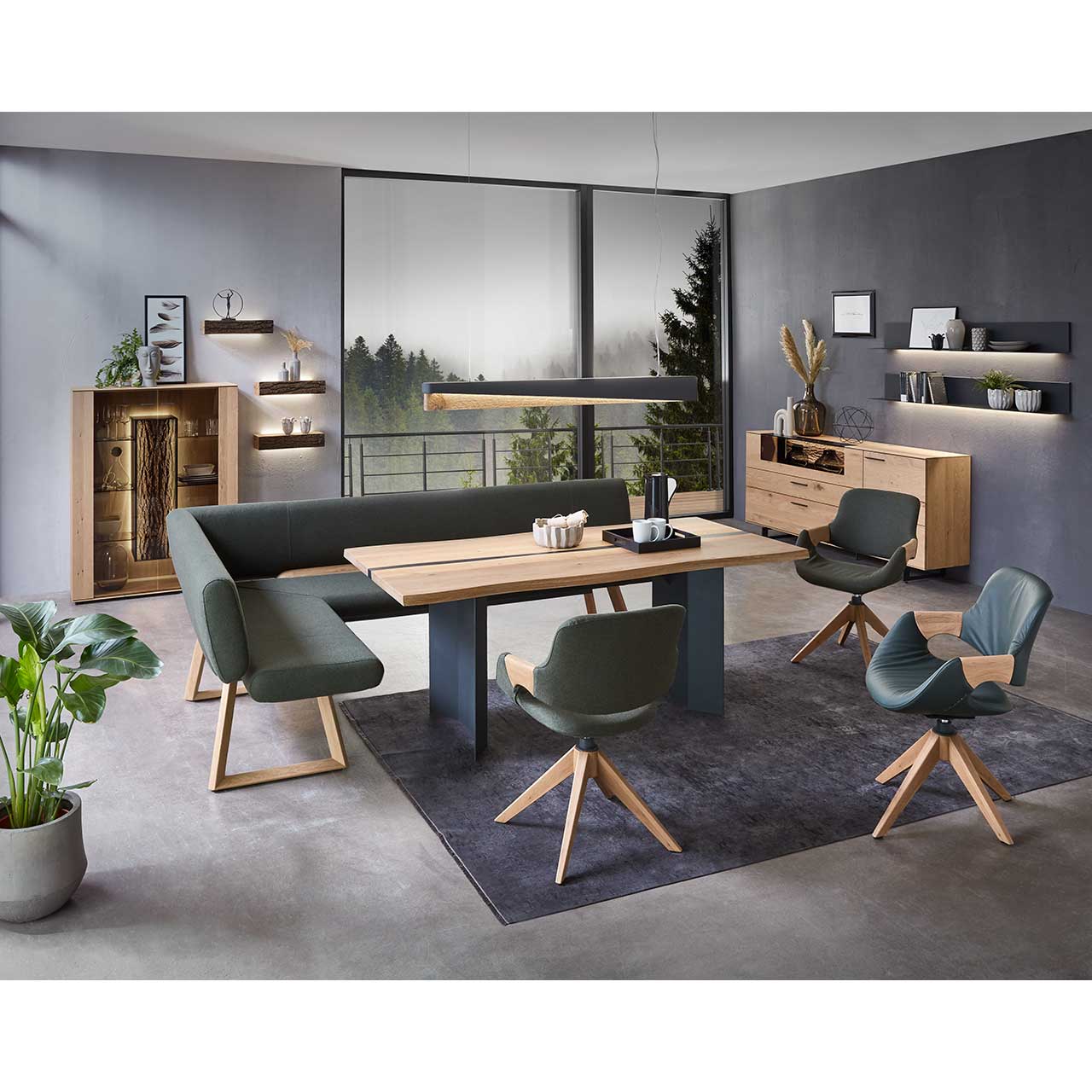 Modernes Esszimmer mit massivem, rechteckigen Holztisch, umgeben von drei Stühlen mit dunkelgrünem Lederbezug auf Holzgestell und der passenden Ecksitzbank. Hinten an der Wand stehend die passenden Esszimmermöbel.
