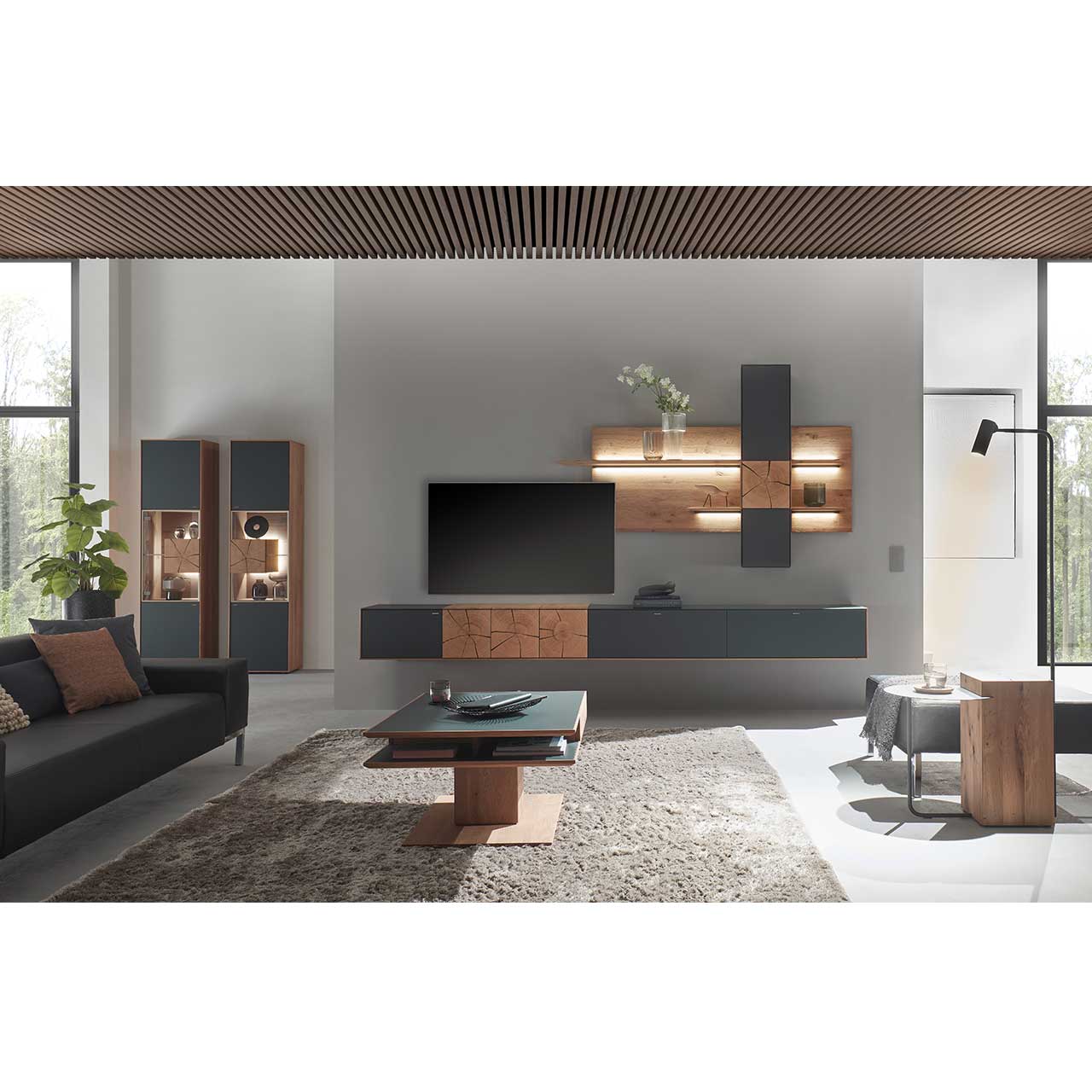 Wohnzimmer mit TV und moderner Wohnwand aus massiver Kerneiche mit Hirnholz und Mattglas anthrazit Fronten, mit Hängelowboard, Wandpaneel mit Regalböden und Hängeschrank. Hinten links zwei passende Standvitrinen. Im Vordergrund rechteckiger Couchtisch.