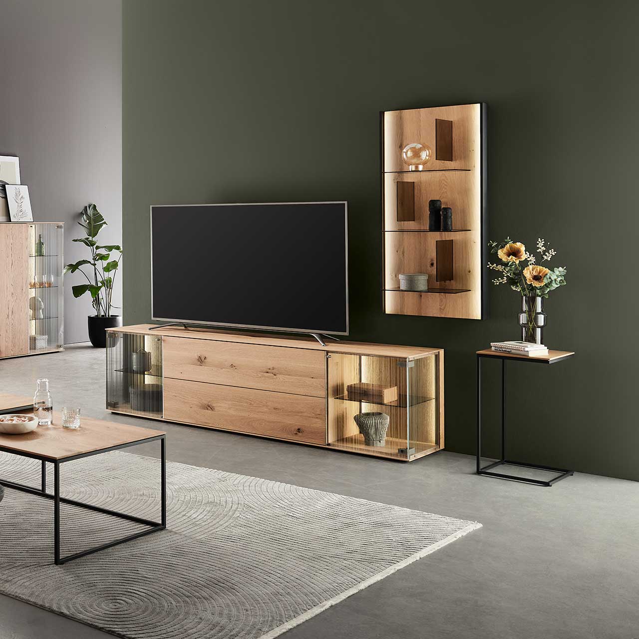 Modernes Wohnzimmer mit zweiteiliger Wohnwand aus Kerneiche Natur: Lowboard mit Glastüren und Wandpaneel mit Glasböden vor dunkelgrauer Wand. Rechts ein passender Beistelltisch, im Vordergrund der dazugehörige Couchtisch.