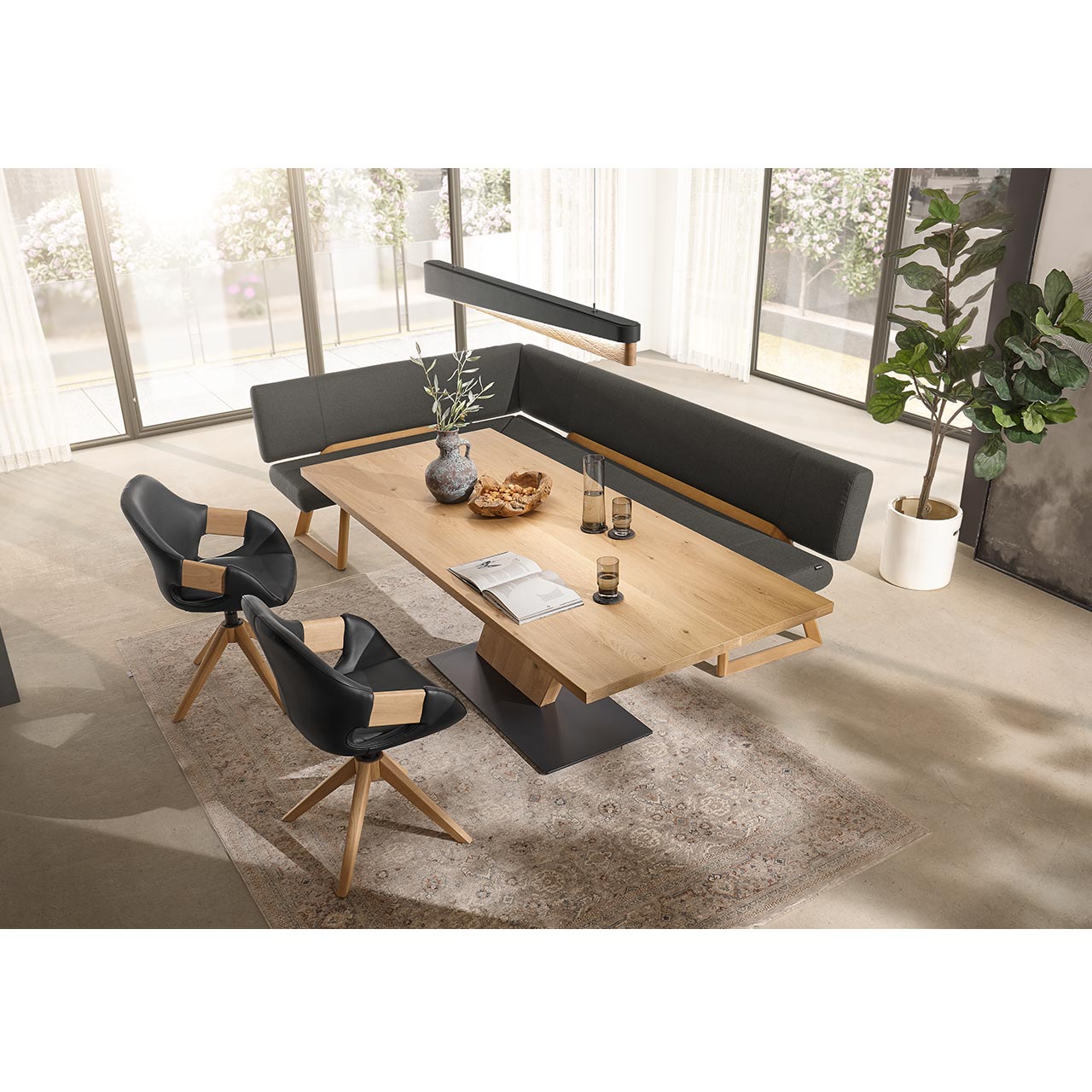 Modernes Esszimmer mit rechteckigem Esstisch aus hellem Holz mit schräg gestelltem Mittelfuß-Element aus Holz und dunkler Bodenplatte. Umgeben von zwei modernen Stühlen und einer Eckbank in dunklem Leder und Holzgestell. Darüber passende Balkenleuchte.