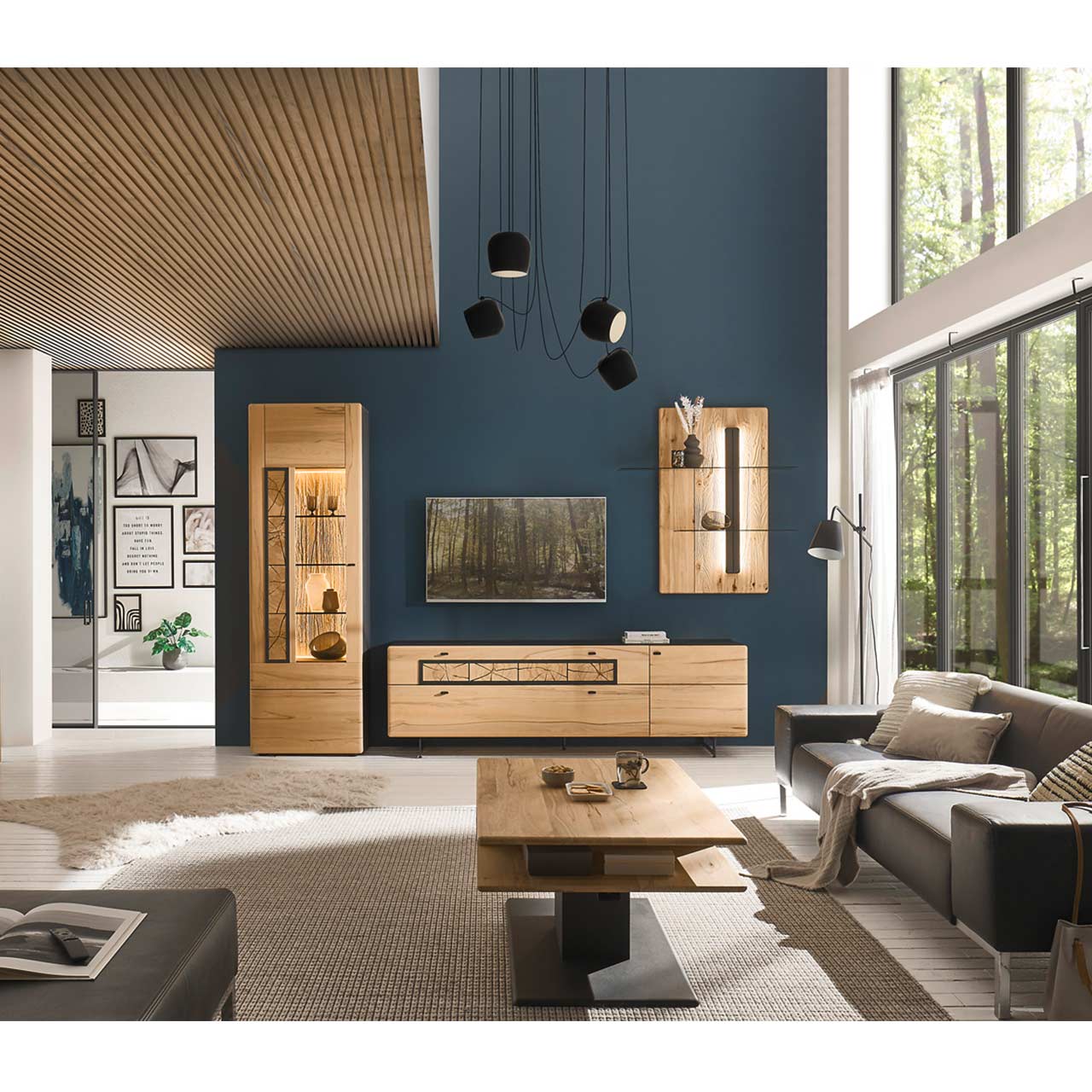 Ambientebild eines modernen Wohnzimmers mit Fernseher, grauer Couch und einer 3teiligen Wohnwand aus Kernbuche bestehend aus Hochvitrine, Lowboard und Wandpaneel. Im Vordergrund steht der passende rechteckige Couchtisch mit Schubkasten und Ablage.