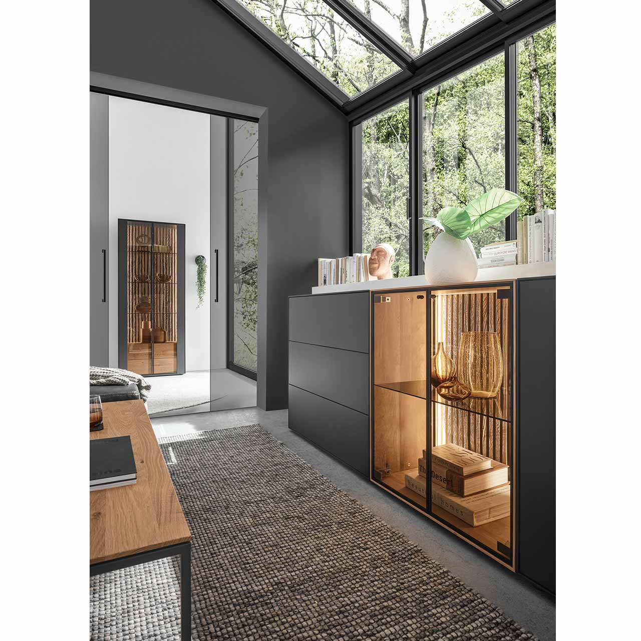 Nahaufnahme der Glastür aus Parsolbronze mit beleuchteter Lamellenrückwand von einem Sideboard aus massiver Birke anthrazit lackiert. Im Hintergrund die passende Standvitrine, links ein minimalistischer Couchtisch.