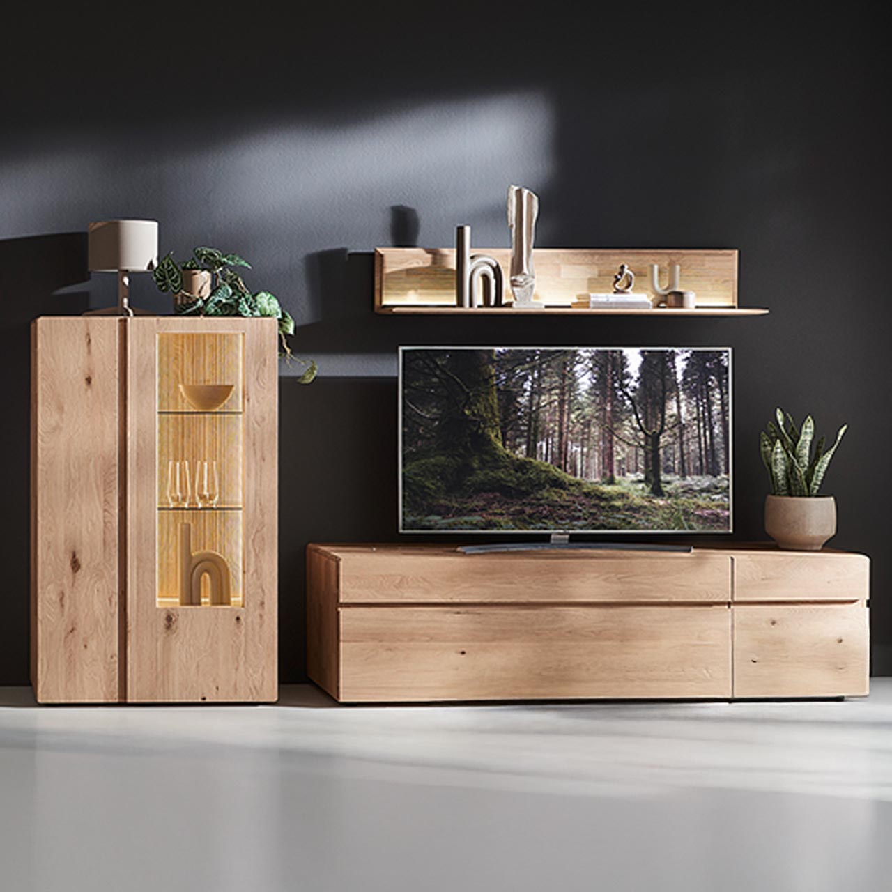 Moderne dreiteilige Wohnwand aus massiver Kerneiche Natur bestehend aus Lowboard mit Tür und Schubkasten, darüber hängend ein Wandpaneel mit Holzablageboden und beleuchteter, geschroppter Rückwand. Links Highboard mit Holz-/Glastüren.