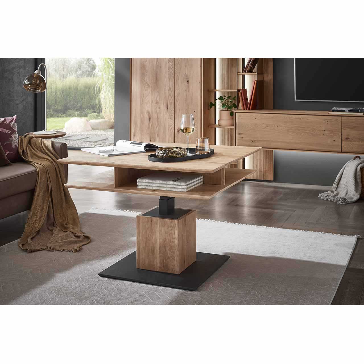Im Wohnzimmer stehender Couchtisch mit quadratischer, höhenverstellbarer Tischplatte aus hellem Eichenholz und zusätzlicher Holzablageplatte, auf massiver, quadratischer Holzsäule und dunkler Bodenplatte. Die Tischplatte ist komplett nach oben gestellt.
