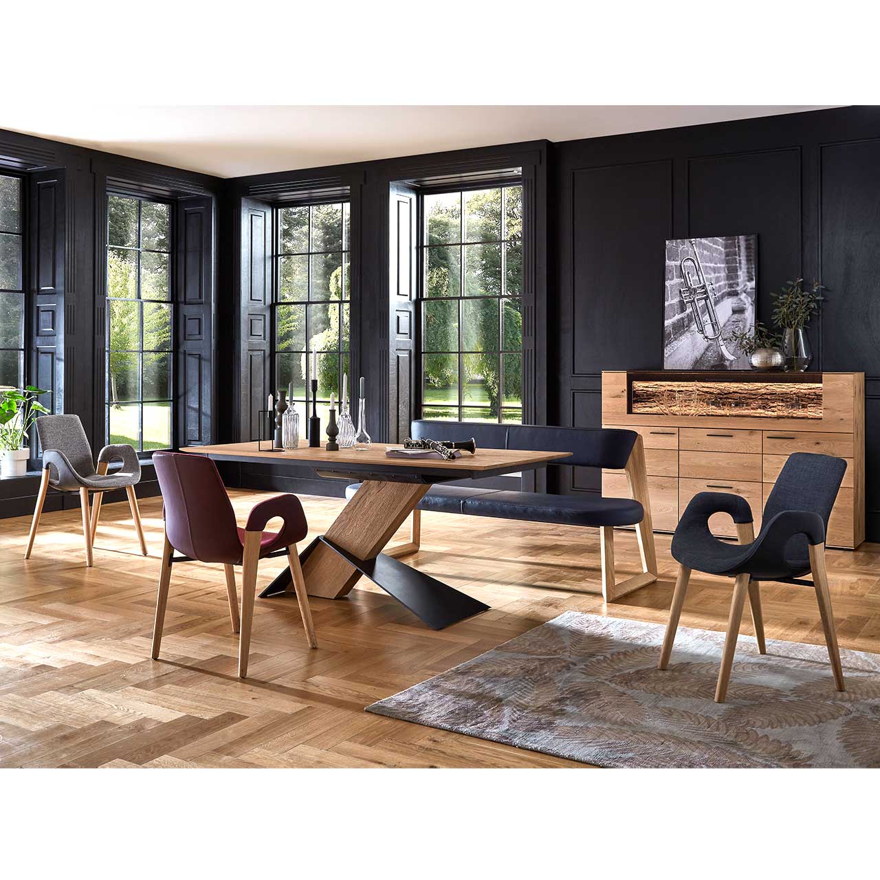 Modernes Esszimmer mit massivem, rechteckigem Holztisch, drei Stühlen auf Holzgestell und einer Sitzbank in schwarzem Leder. Dahinter vor dunkler Wand das passende Highboard mit Glasausschnitt.