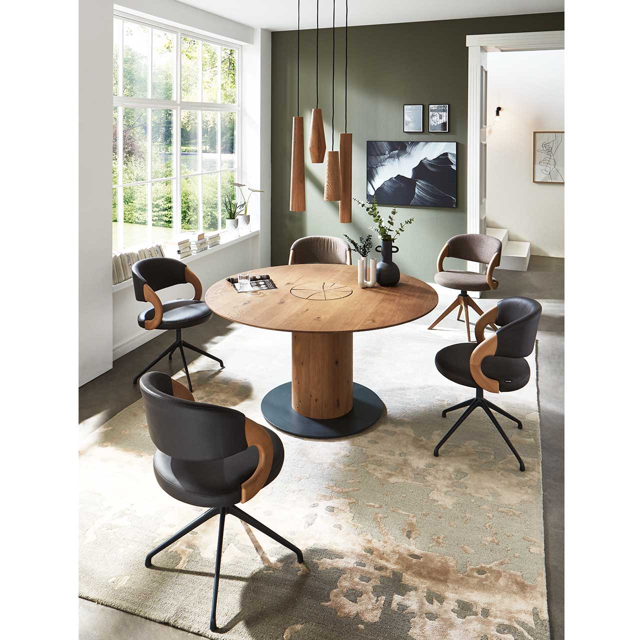 Modernes Esszimmer mit massivem, runden Holztisch, umgeben von fünf Stühlen mit dunklem Bezug auf Metallgestell. Über dem Tisch hängen vier Pendelleuchten mit zylinderförmigen Lampenschirmen aus Holz.