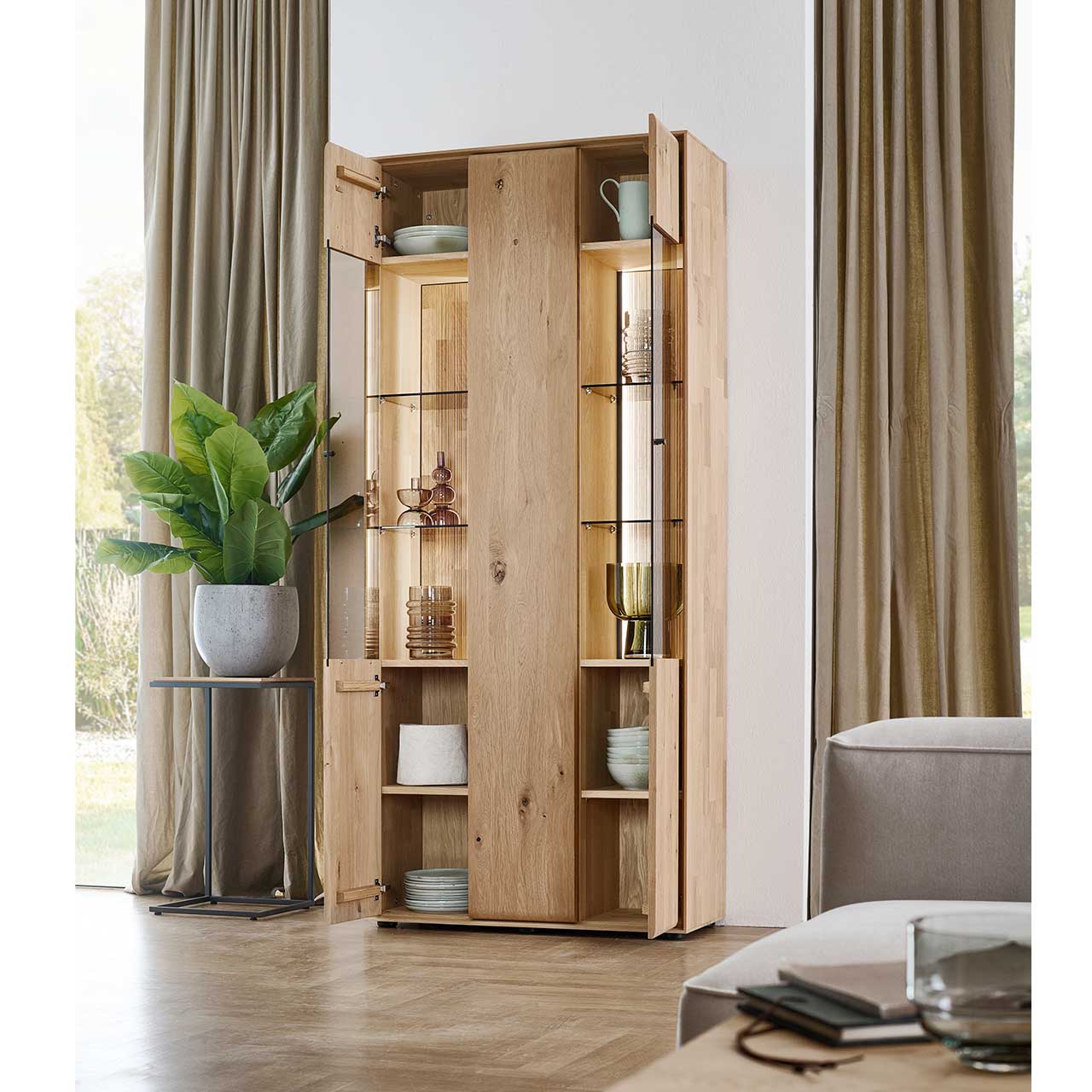 Moderne Standvitrine aus massivem Eichenholz mit 2 geöffneten Glastüren rechts und links und einer geschlossenen Holztür mittig - im Wohnzimmer stehend.