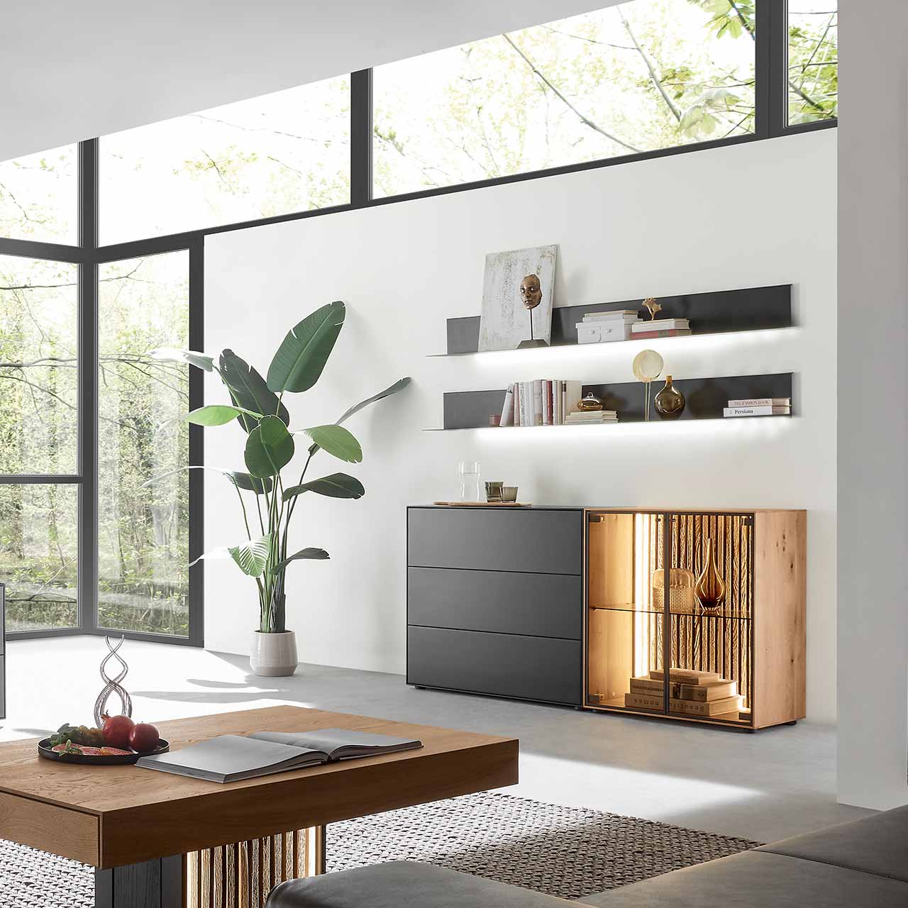 Modernes Wohnzimmer mit beleuchtetem Sideboard aus Birke anthrazit mit Glastüren, Lamellenrückwand und dunklen Schubkästen, darüber hängend zwei Metallregale in anthrazit. Im Vordergrund der passende Holz-Couchtisch.