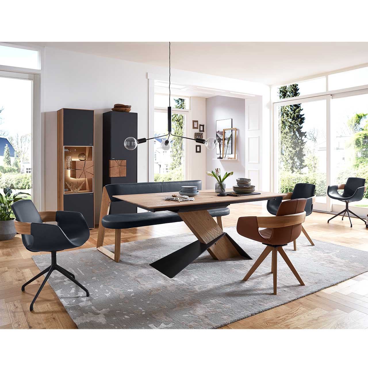 Modernes Esszimmer mit massivem, rechteckigem Holztisch, drei Stühlen auf Holzgestell sowie Metallgestell und einer Sitzbank in schwarzem Leder. Dahinter an der Wand eine passende Vitrine und ein Hochschrank mit Hirnholzscheiben.