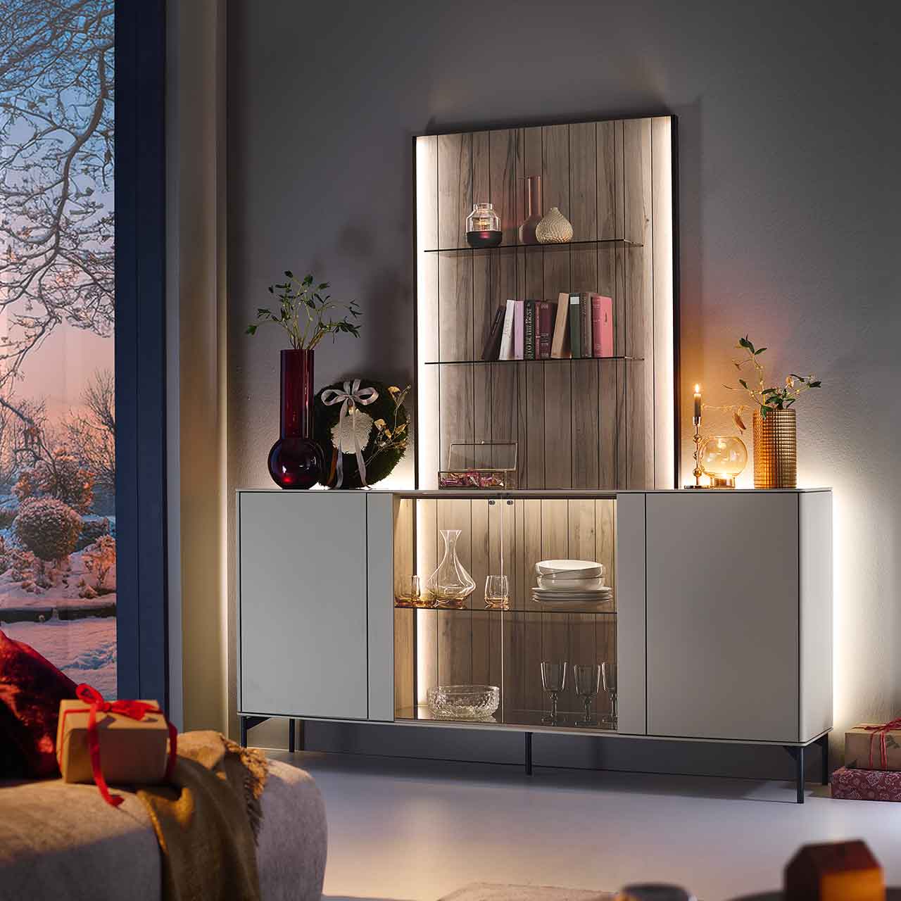 Modernes Massivholz-Sideboard auf Metallfüßen mit zwei Türen rechts und links lackiert in Sandfarben, mittig mit Glasausschnitt und Glasboden vor beleuchteter Holzrückwand in edler, dunkelbrauner Altholzbuche Uyaa, darüber Holzpaneel mit Glasböden.