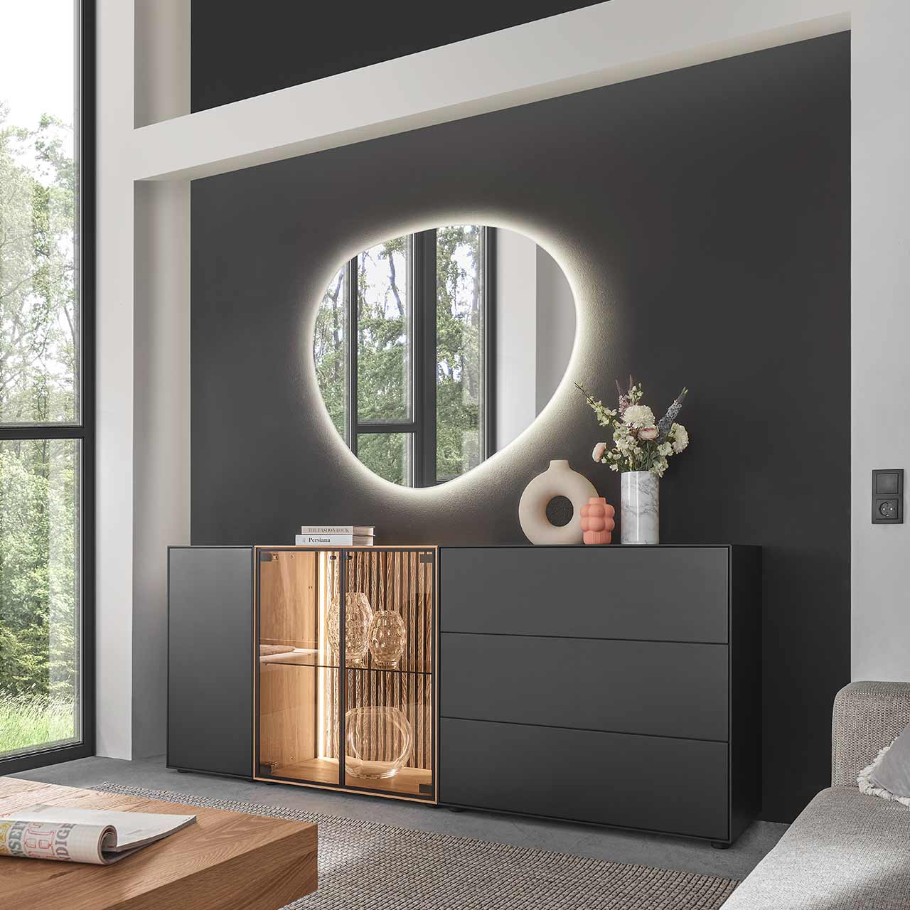 Modernes Sideboard aus massiver Birke anthrazit mit Holztür, zwei Glastüren, beleuchteter heller Holz-Lamellenrückwand und Glaseinlegeboden sowie zwei Schubkästen und Klappe in Anthrazit. Darüber hängt ein asymmetrischer Spiegel - vor dunkelgrauer Wand.