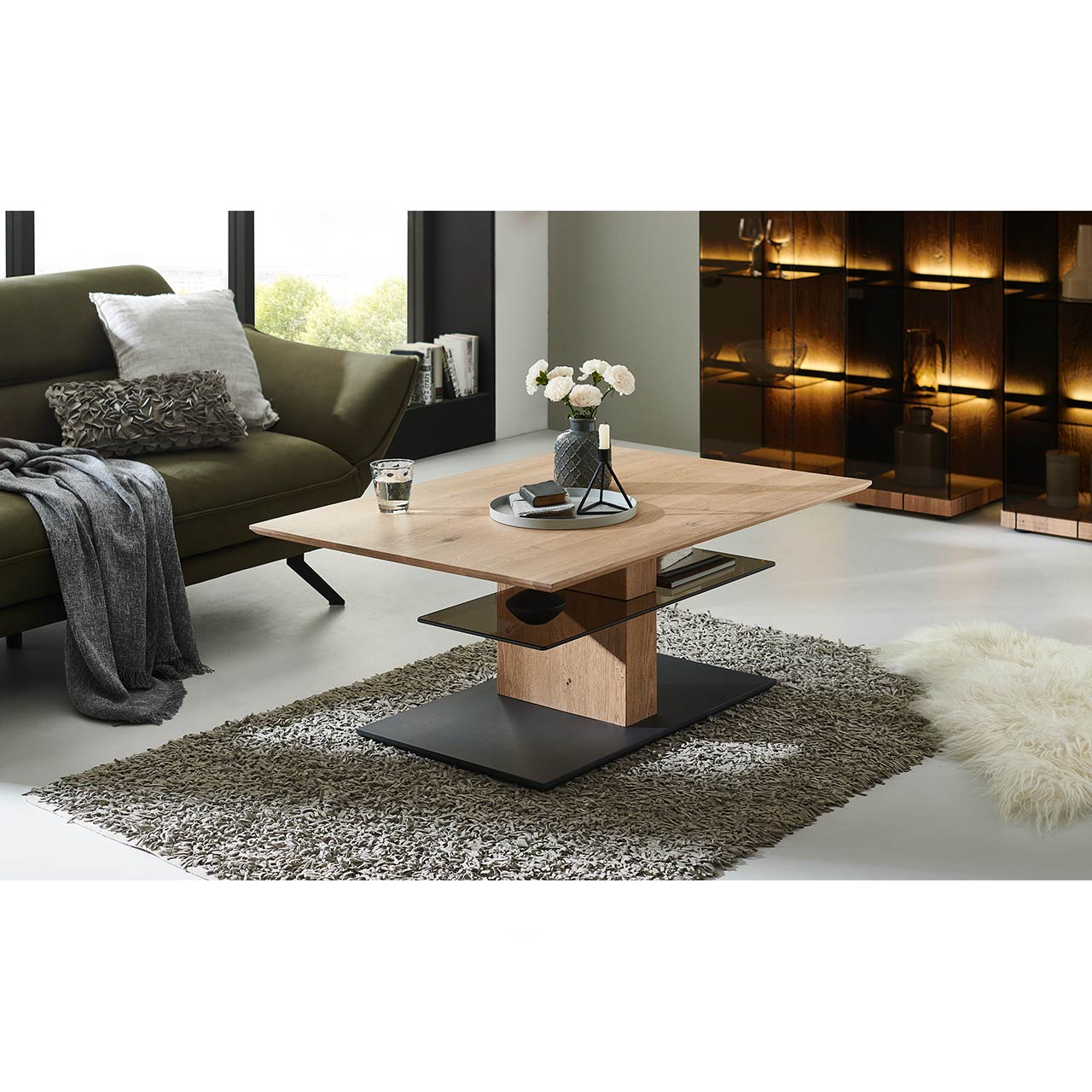Milieubild Wohnzimmer - Hartmann NATURWERKE - Couchtisch 8400C-1424 Im Wohnzimmer stehender Couchtisch mit rechteckiger, höhenverstellbarer Tischplatte aus Holz und zusätzlicher fester Glasablage darunter - auf massiver Holzsäule und dunkler Bodenplatte.