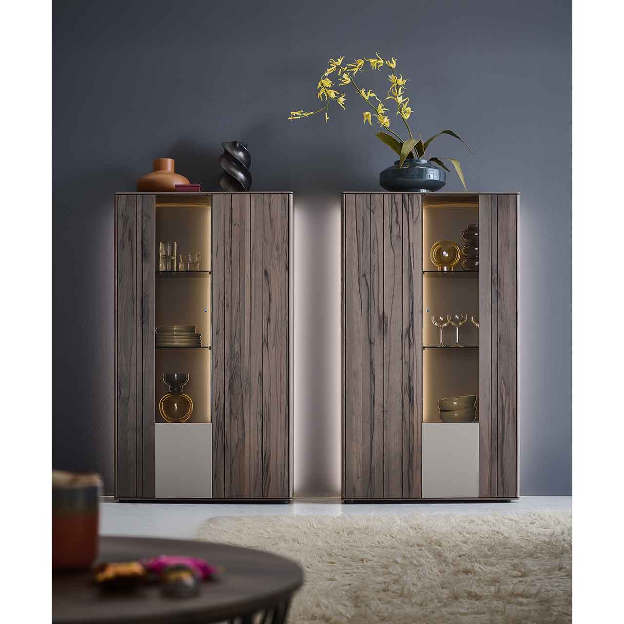 Milieubild - Hartmann AUDRA - Highboards 5610-8095 und 8096 - Altholzbuche Uyaa Wohnzimmer mit zwei modernen Highboards aus massiver, dunkelbrauner Altholzbuche Uyaa, jeweils mit beleuchtetem Glasausschnitt und Glasböden, vor beleuchteter Rückwand in lack Sand.