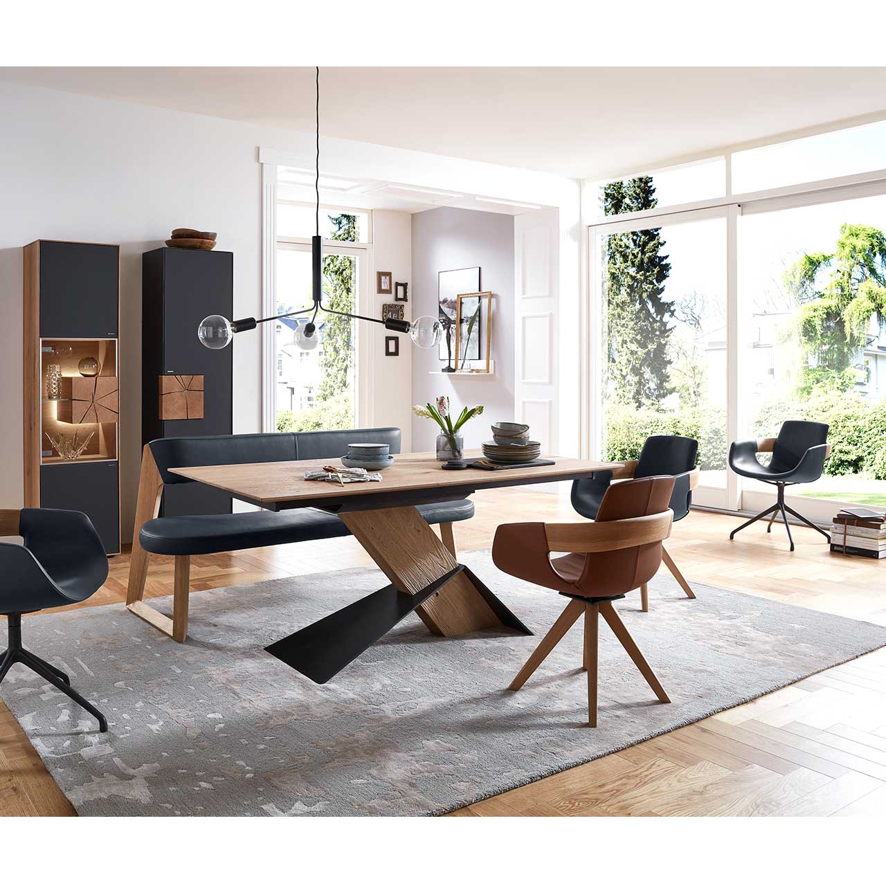 Modernes Esszimmer mit massivem, rechteckigem Holztisch, drei Stühlen auf Holzgestell sowie Metallgestell und einer Sitzbank in schwarzem Leder. Dahinter an der Wand eine passende Vitrine und ein Hochschrank mit Hirnholzscheiben.
