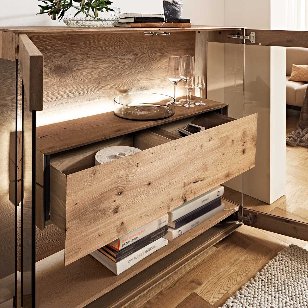 Geöffneter Doppelschubkasten im beleuchteten Hirnholzblock des Highboards CAYA 7170-HM-6139, mit Einblick in den Stauraum hinter der Parsolbronze-Glastür