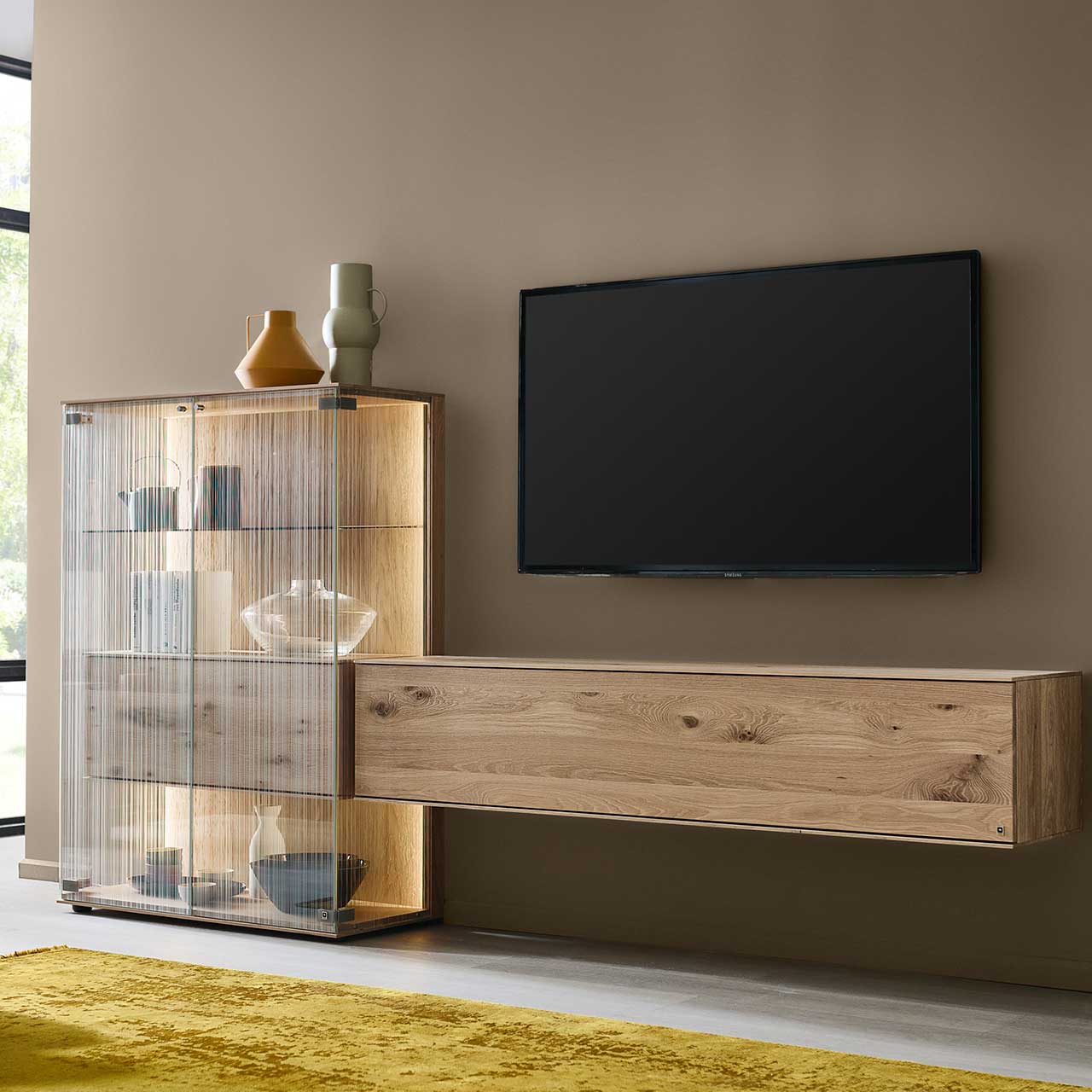 LINEA Leonardo Living - Wohnwand 8540 Nr. V24 Modernes Wohnzimmer mit Fernseher und zweiteiliger Wohnwand aus Kerneiche Natur: Highboard mit Glastüren aus satiniertem Glas und innenliegendem Holzkorpus mit Klappe und einem schlichten Hängelowboard – platziert vor einer dunklen Wand.