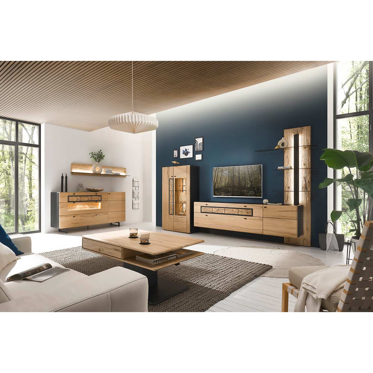 Eingerichtetes Wohnzimmer mit moderner Wohnwand aus Kernbuche vor einer dunkelblauen Wand bestehend aus Paneel, Lowboard und Highboard. Rechts das passende Sideboard auf Metallkufen darüber ein Regal. Im Vordergrund der dazugehörige Couchtisch.