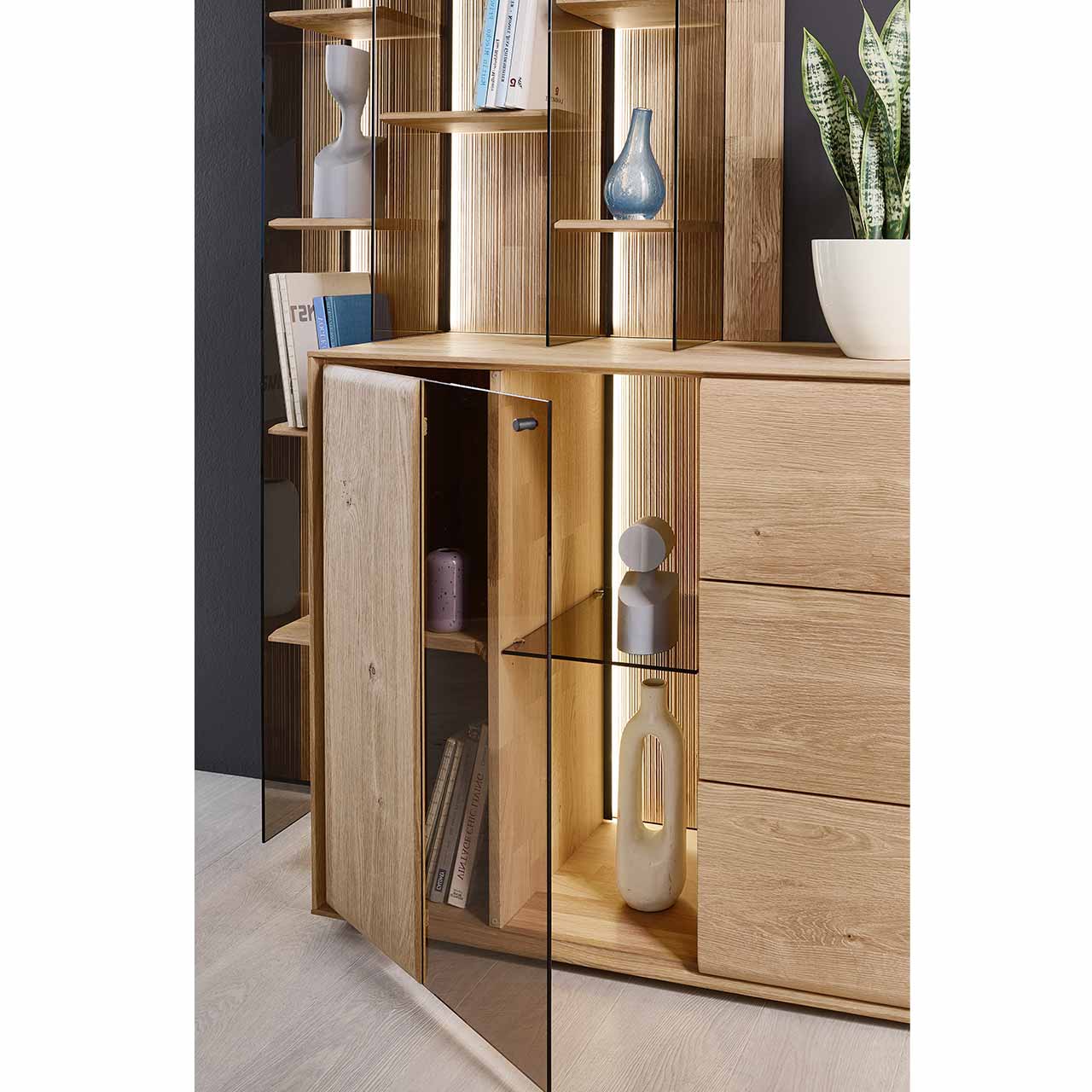 Detailaufnahme eines Sideboard aus massiver Kerneiche Natur mit beleuchteter und geöffneter Glas/Holztür, innenliegend sichtbar die geschoppte Rückwand, ein Glaseinlegeboden und ein Holzboden. Hinter passendes Wandregal als Anbauelement.