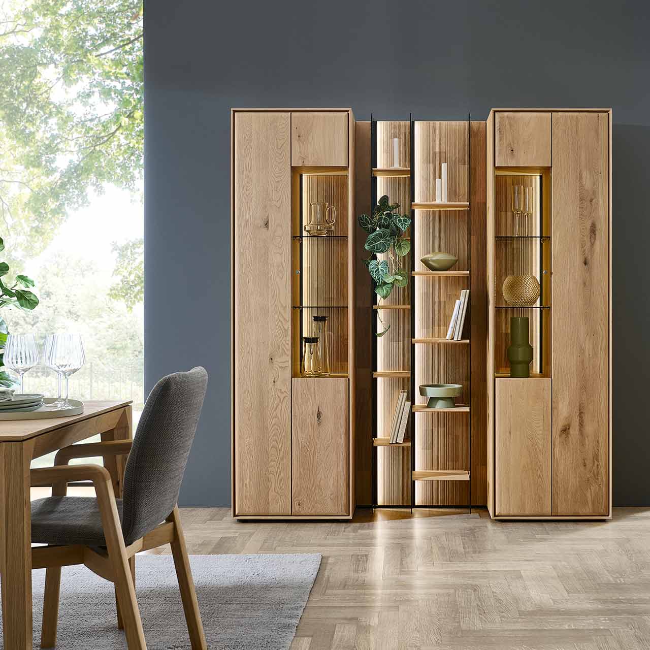 Zwei moderne Standvitrinen aus massivem Eichenholz mit je einer Holztür und einer Glas/Holztür, mittig dazwischen steht ein hohes Wandregal mit beleuchteten Holz und Glasböden - vor dunkelgrauer Wand im Wohnzimmer stehend.