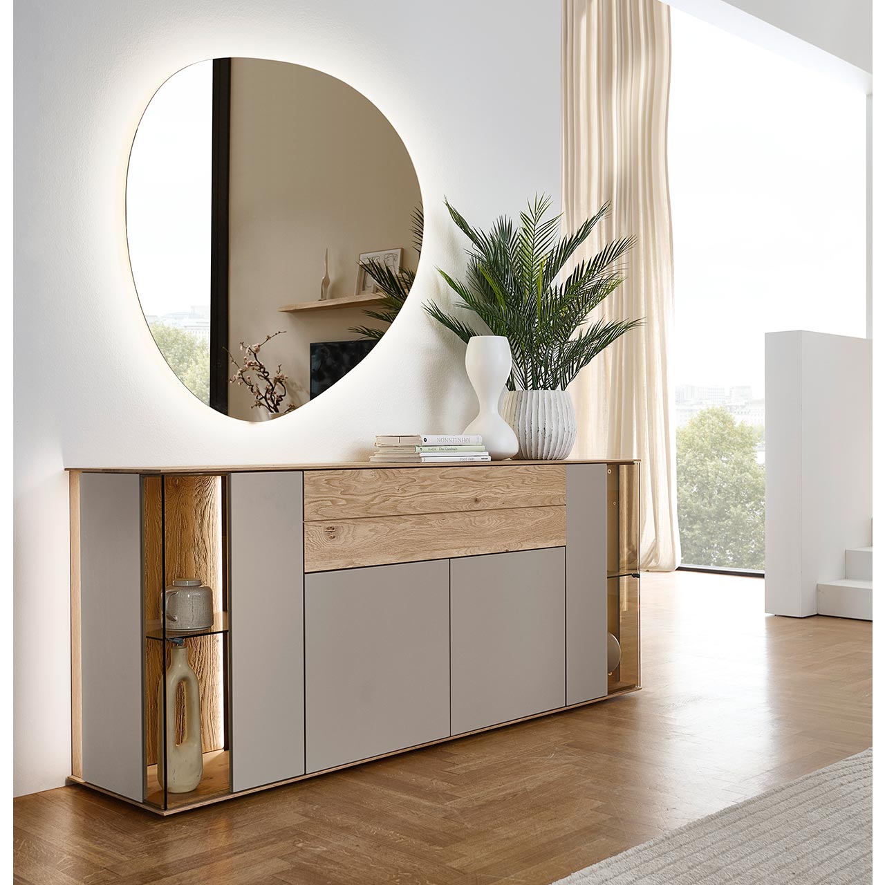 Hartmann Möbel SVEA - Sideboard 8660-4191 - Kerneiche Natur, Lack Sand - Milieubild Modernes Sideboard aus massiver Kerneiche Natur mit Lackfronten in Sand und Riffholz. Mit zwei Lacktüren unten, einem langen Schubkasten mit Riffholzfront oben und je einer Glas-Lacktür rechts und links mit beleuchteter Riffholzrückwand - in Frontansicht.