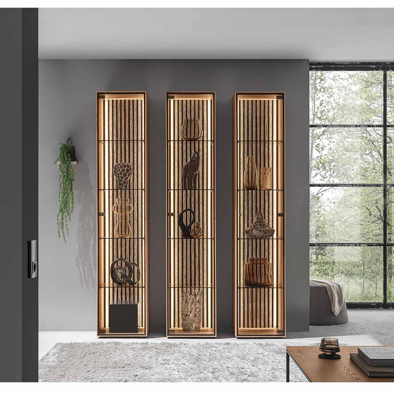 Drei beleuchtete Standvitrinen aus hellem Massiv-Holz mit Lamellenrückwand und je vier Glaseinlegeböden - vor hellgrauer Wand.
