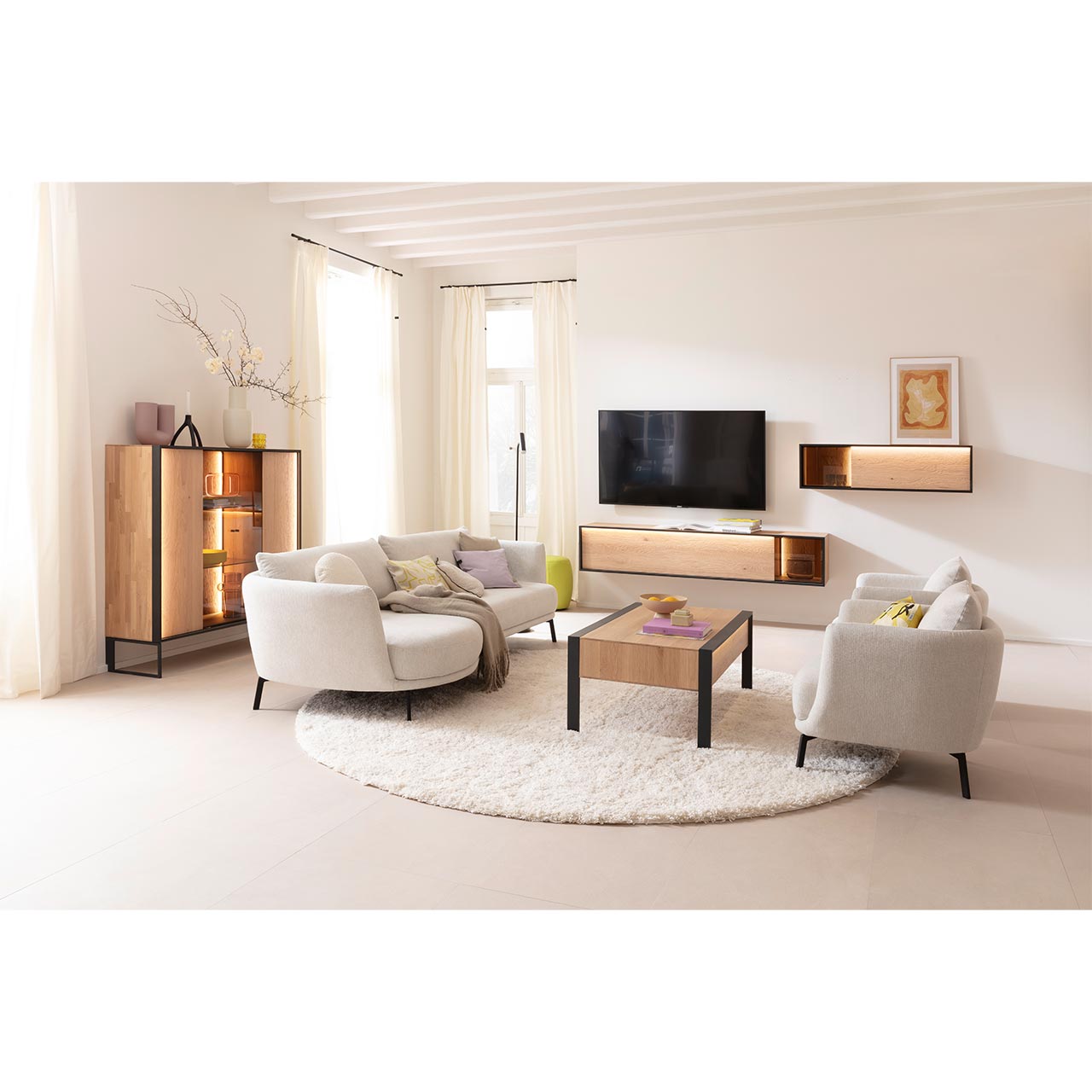 Eingerichtetes Wohnzimmer mit Massivholzmöbeln aus heller Kerneiche Natur: Highboard auf Metallkufen anthrazit mit beleuchteter Front und Glastüren, schlichtes Hängelowboard, Hängeschrank mit Glasausschnitt und passender Holz-Couchtisch auf Metallfüßen. 