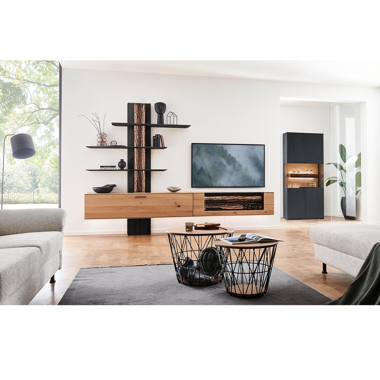 Wohnzimmer mit Wohnwand aus zwei Hängelowboards in massiver Kerneiche Natur, teils verglast, einem Wandpaneel mit beleuchteter Rinden-Rückwand und drei langen Ablageböden, dazu rechts eine Standvitrine in anthrazit gebeizter Kerneiche.