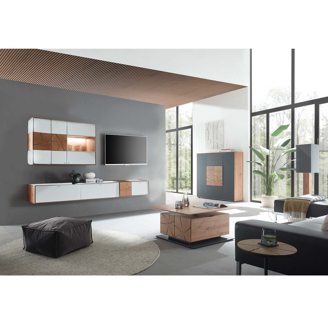 Wohnzimmer mit TV und moderner Wohnwand aus massiver Kerneiche und Mattglas weiß Fronten, mit schmalem Hängelowboard und beleuchteter Hängevitrine vor dunkelgrauer Wand. Rechts das passende Highboard in Anthrazit. Im Vordergrund Couchtisch mit Glasplatte.