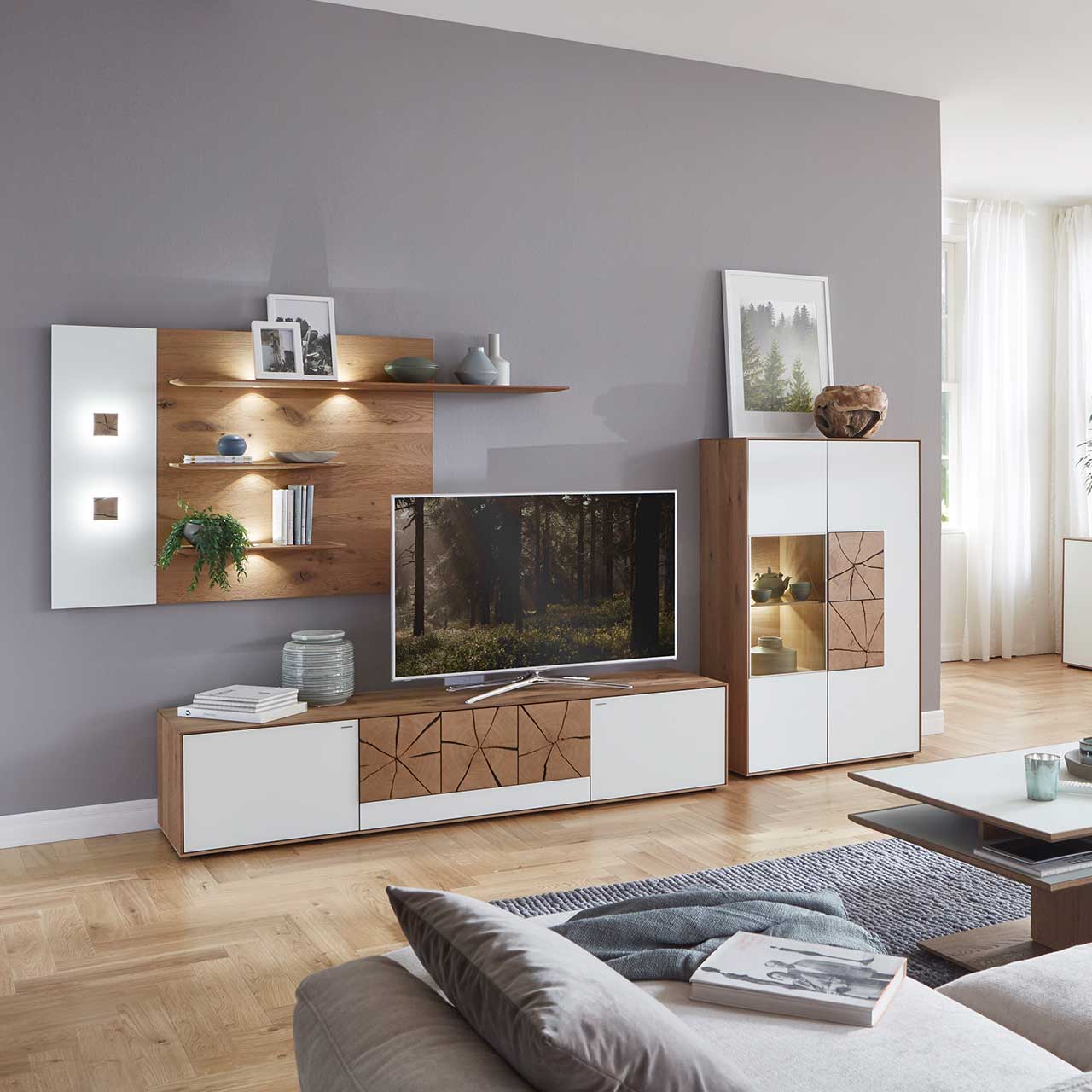 Milieubild Hartmann CAYA - Standelement 7170-7096W re. Wohnzimmer mit moderner Massivholz Wohnwand mit Mattglas weiß Fronten und dekorativen Hirnholzscheiben: Langes Lowboard mit Hirnholz, Wandpaneel mit zwei Regalböden und Highboard mit beleuchtetem Glasausschnitt und Hirnholz.