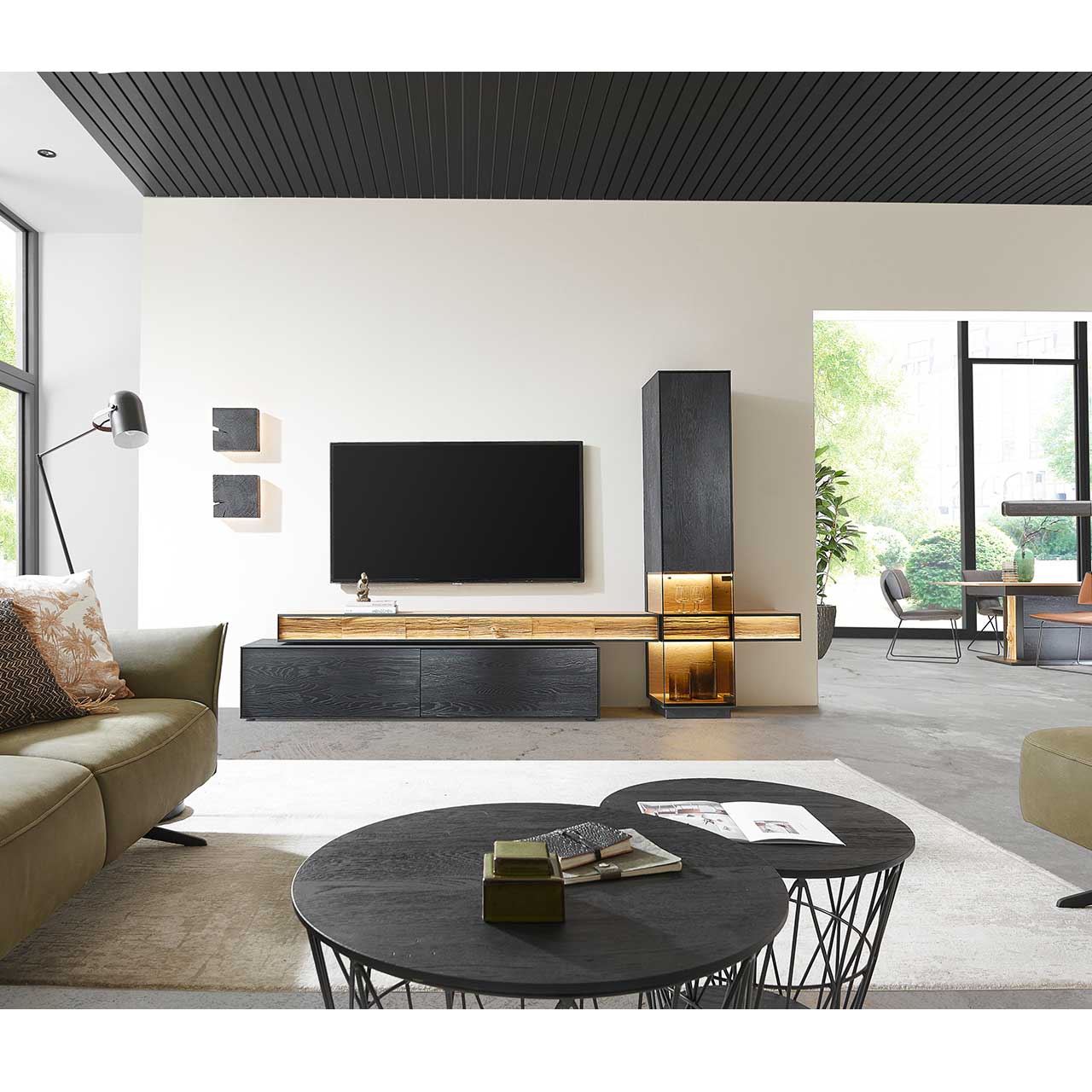 Wohnzimmer mit moderner Wohnwand aus massiver Kernesche anthrazit: Lowboard, Standvitrine und zwei TV-Brücken. Im Vordergrund stehen zwei passende Couchtische mit Metallkorb. 